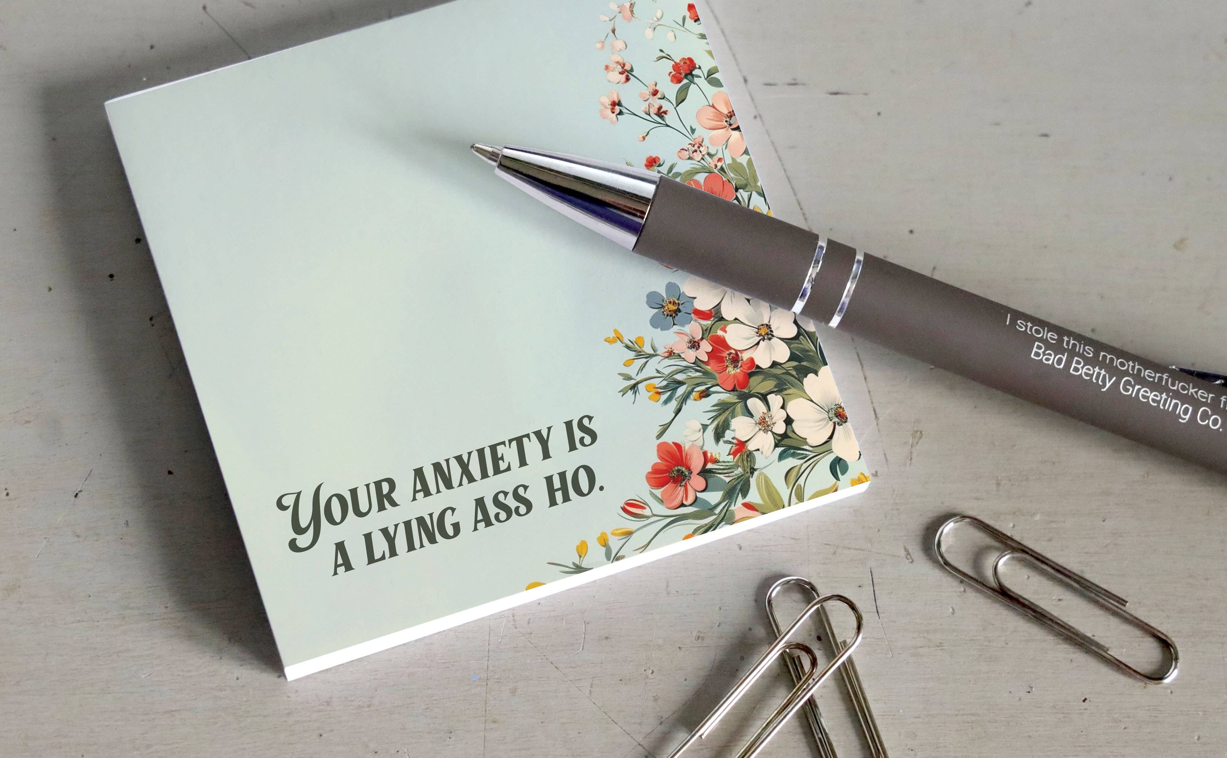 Notebook with a floral design and humorous text on a desk with a pen and paper clips.