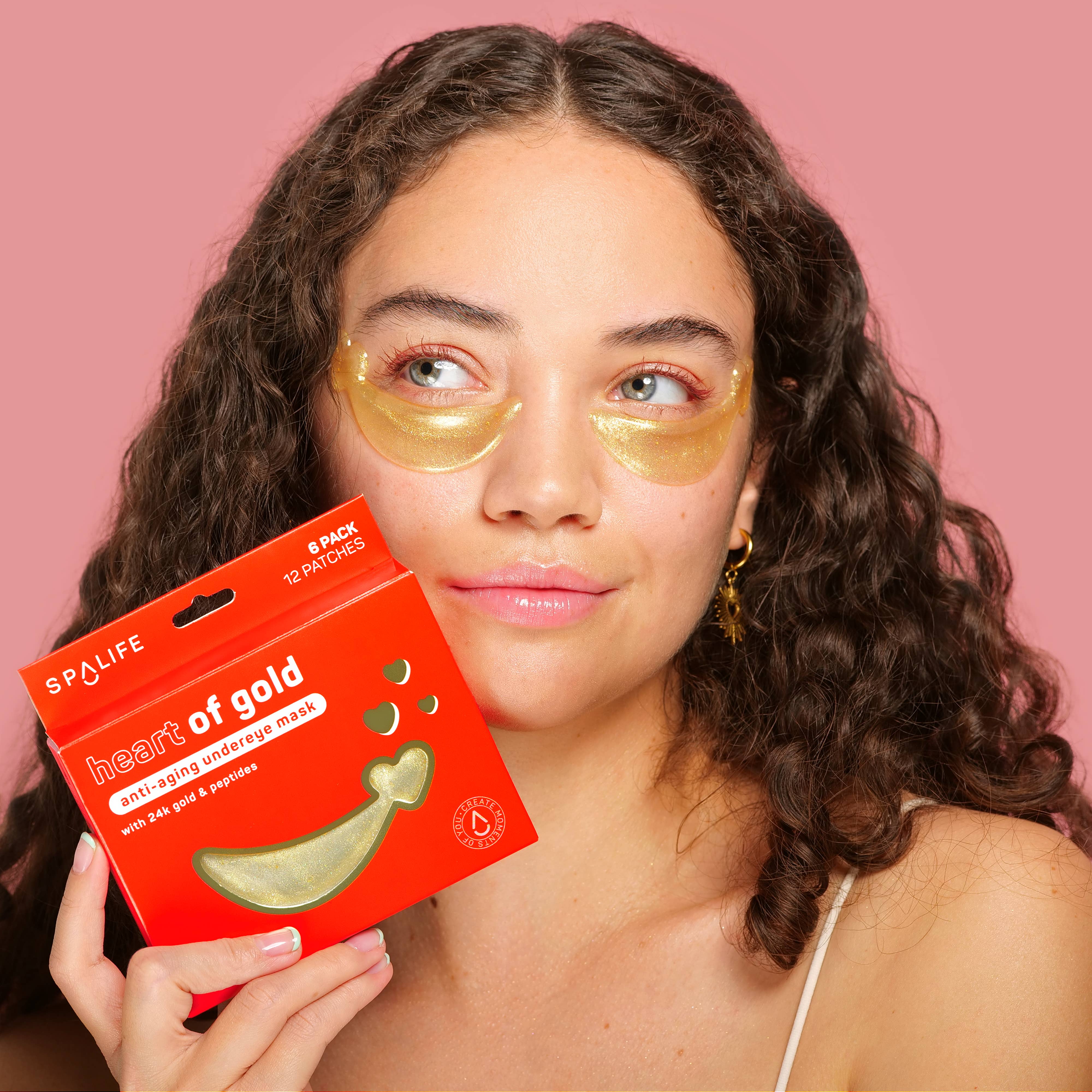 A woman with gold under-eye masks holds a red "heart of gold" eye mask box, smiling slightly. The background is a soft pink, conveying a relaxed tone.