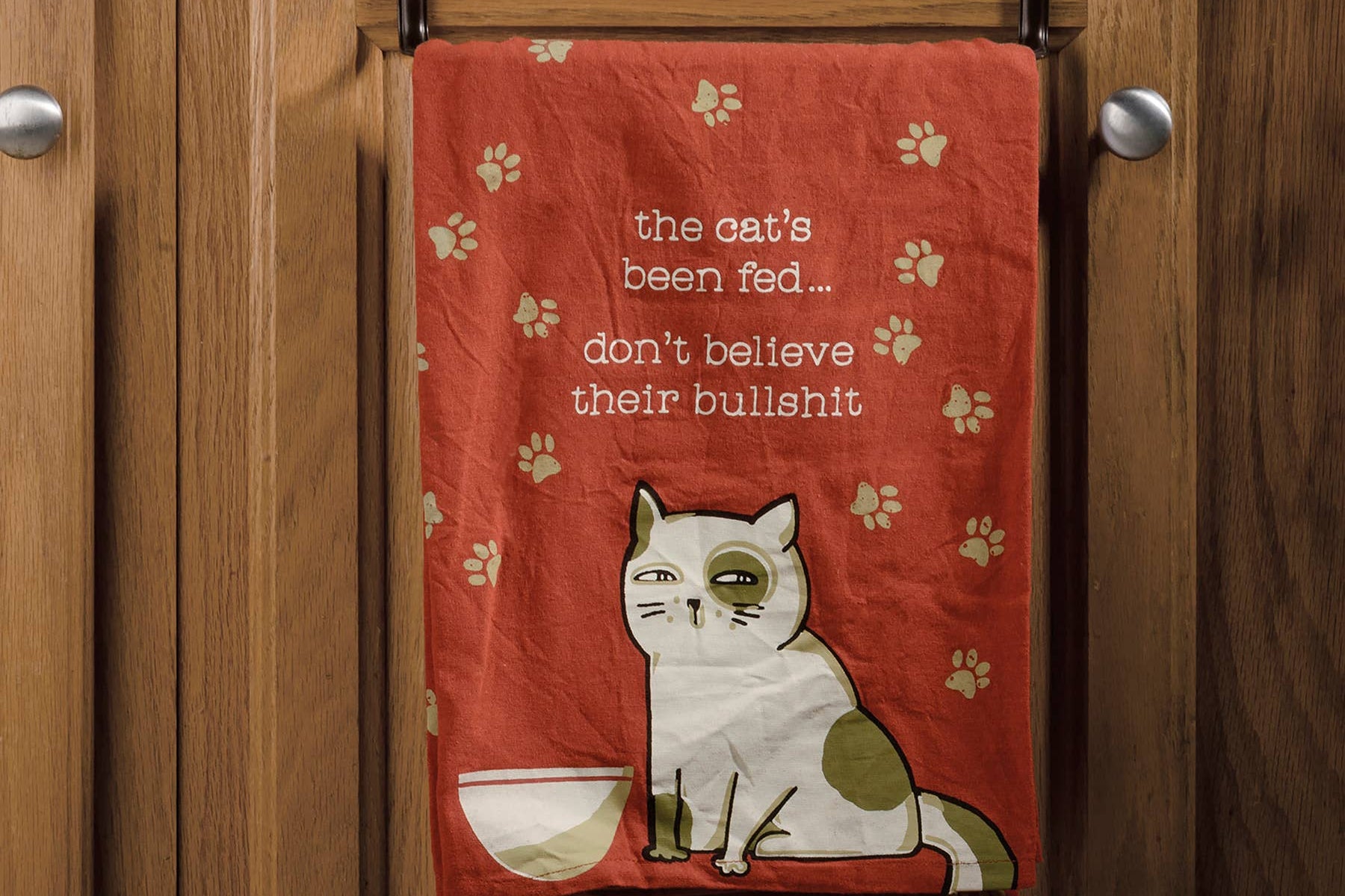 A red dish towel with paw prints features a grumpy cat illustration beside an empty bowl. Text reads, "The cat's been fed... don't believe their bullshit," creating a playful tone.