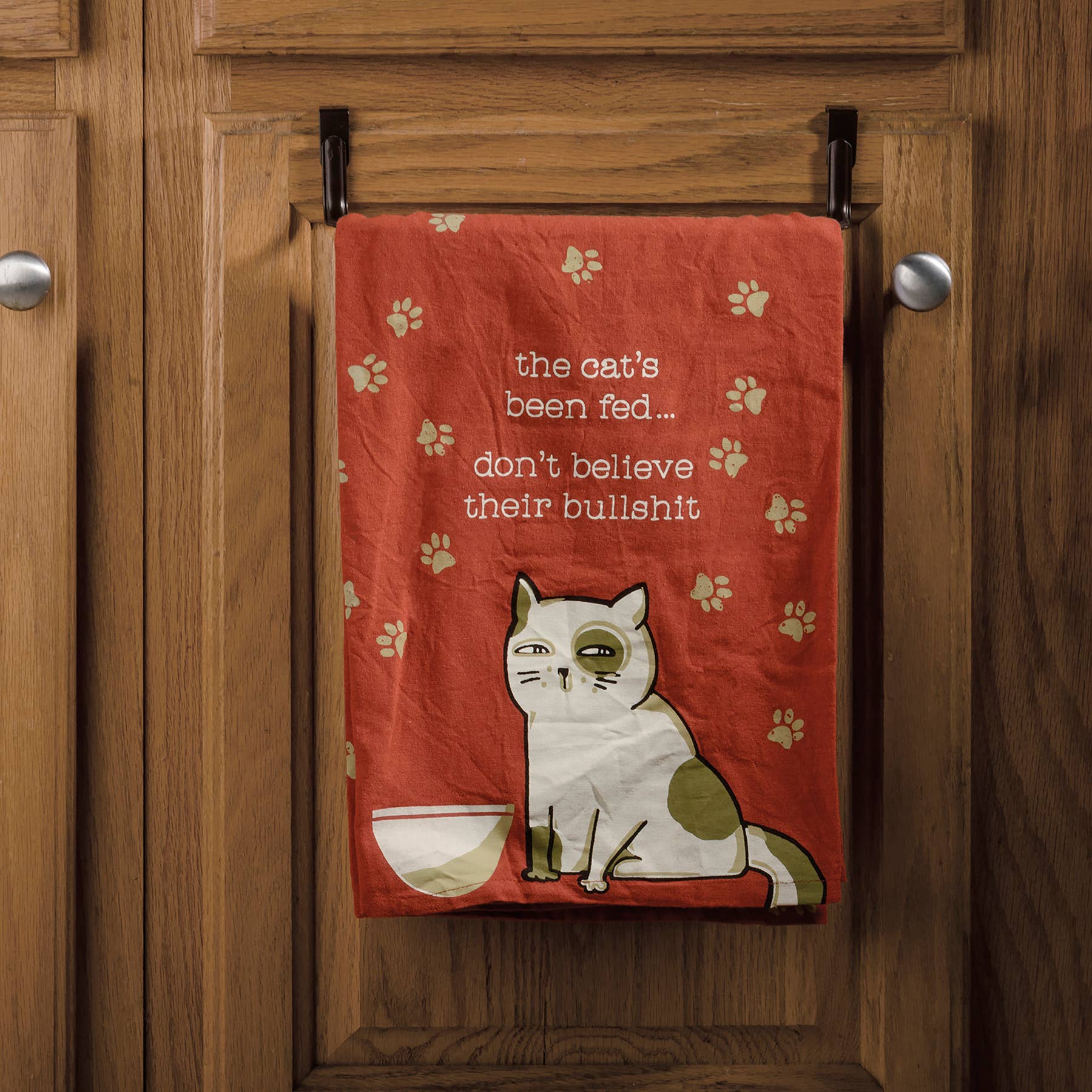A red dish towel with paw prints features a grumpy cat illustration beside an empty bowl. Text reads, "The cat's been fed... don't believe their bullshit," creating a playful tone.