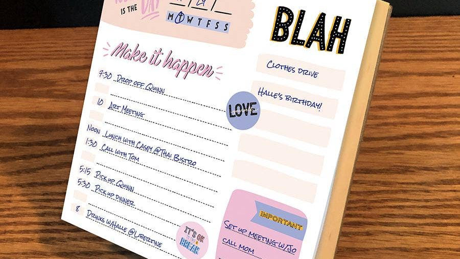 A colorful desk planner page on a wooden surface displays a daily schedule with handwritten tasks. Words like "Blah" and "Love" add a playful tone.