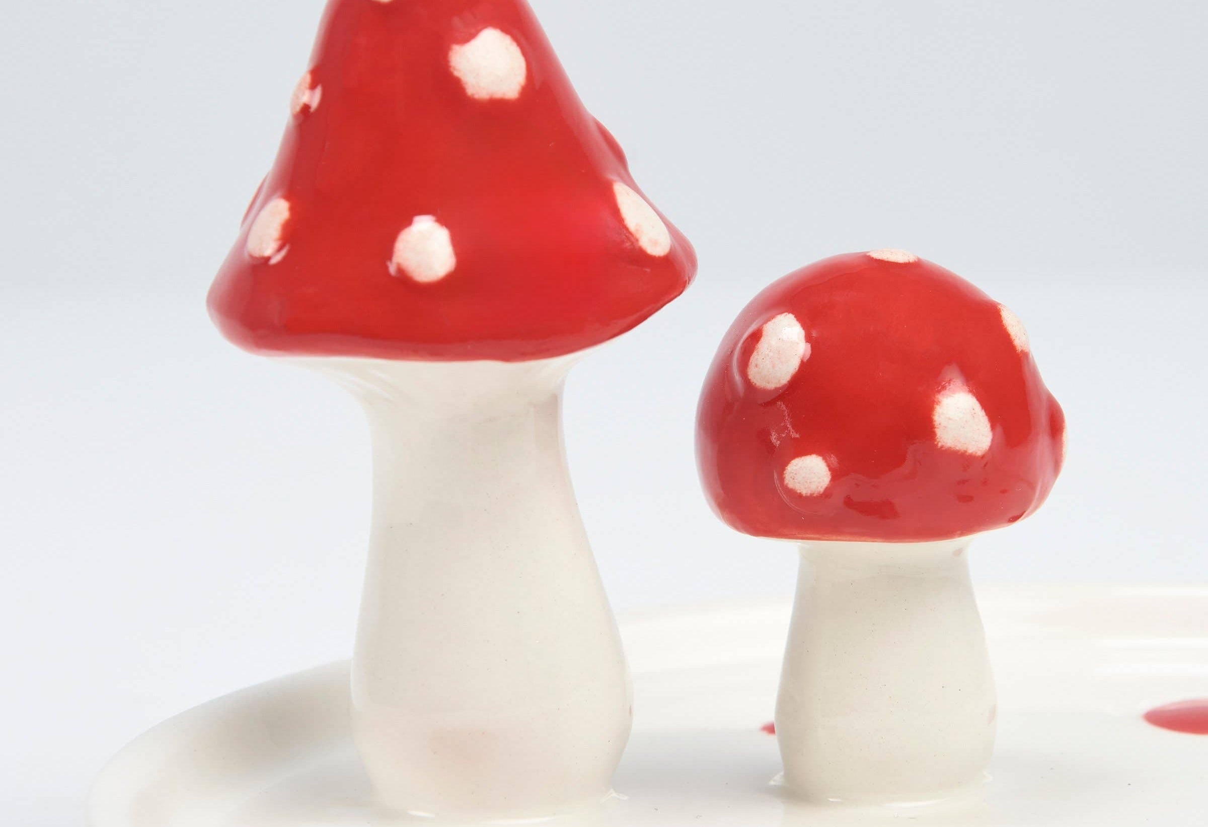 Two ceramic mushrooms with glossy red caps and white spots stand on a white base. The whimsical tones add a playful, decorative touch.