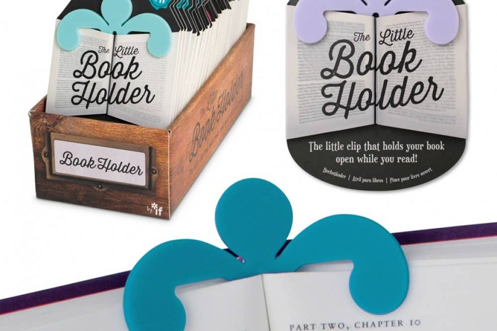 A colorful book holder clip is shown in use, holding a book open. The package has decorative designs and text reading "The Little Book Holder." Display box is visible.