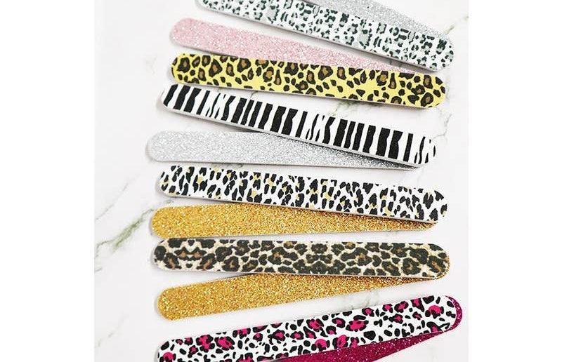 An assortment of ten glittery and patterned emery board nail files fanned out on a white marble background, featuring zebra stripes, cheetah print, and hot pink leopard print.
