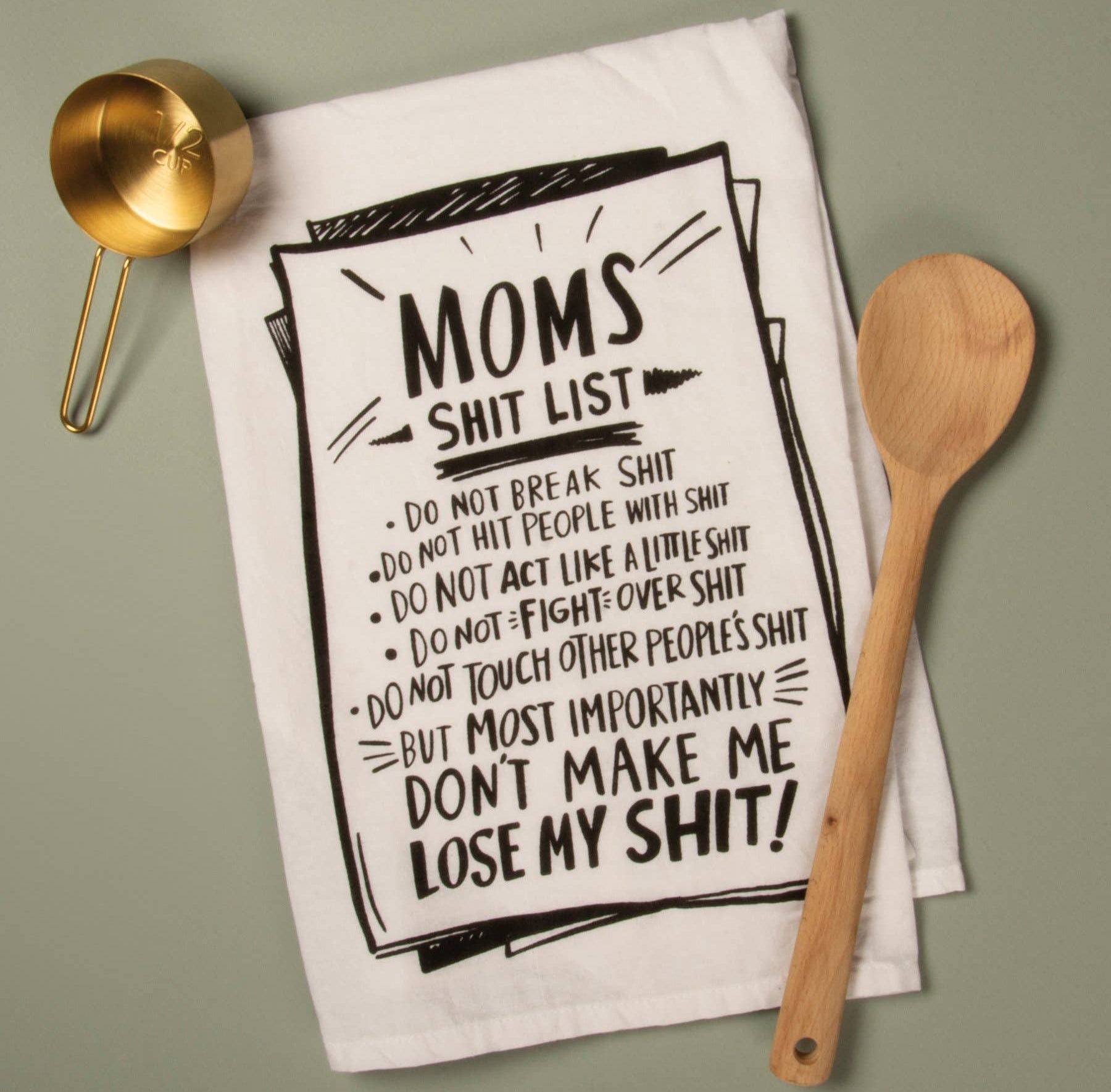 A humorous kitchen towel featuring a list titled "Mom's Shit List" with comedic rules written in bold black text. A gold measuring cup and wooden spoon are placed beside it. The tone is playful and lighthearted.