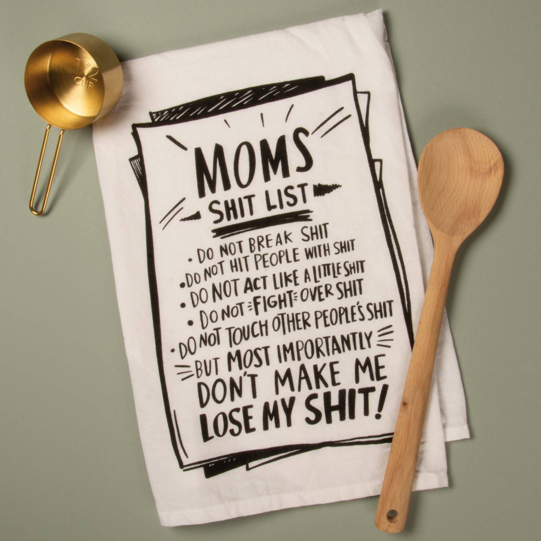 A humorous kitchen towel featuring a list titled "Mom's Shit List" with comedic rules written in bold black text. A gold measuring cup and wooden spoon are placed beside it. The tone is playful and lighthearted.