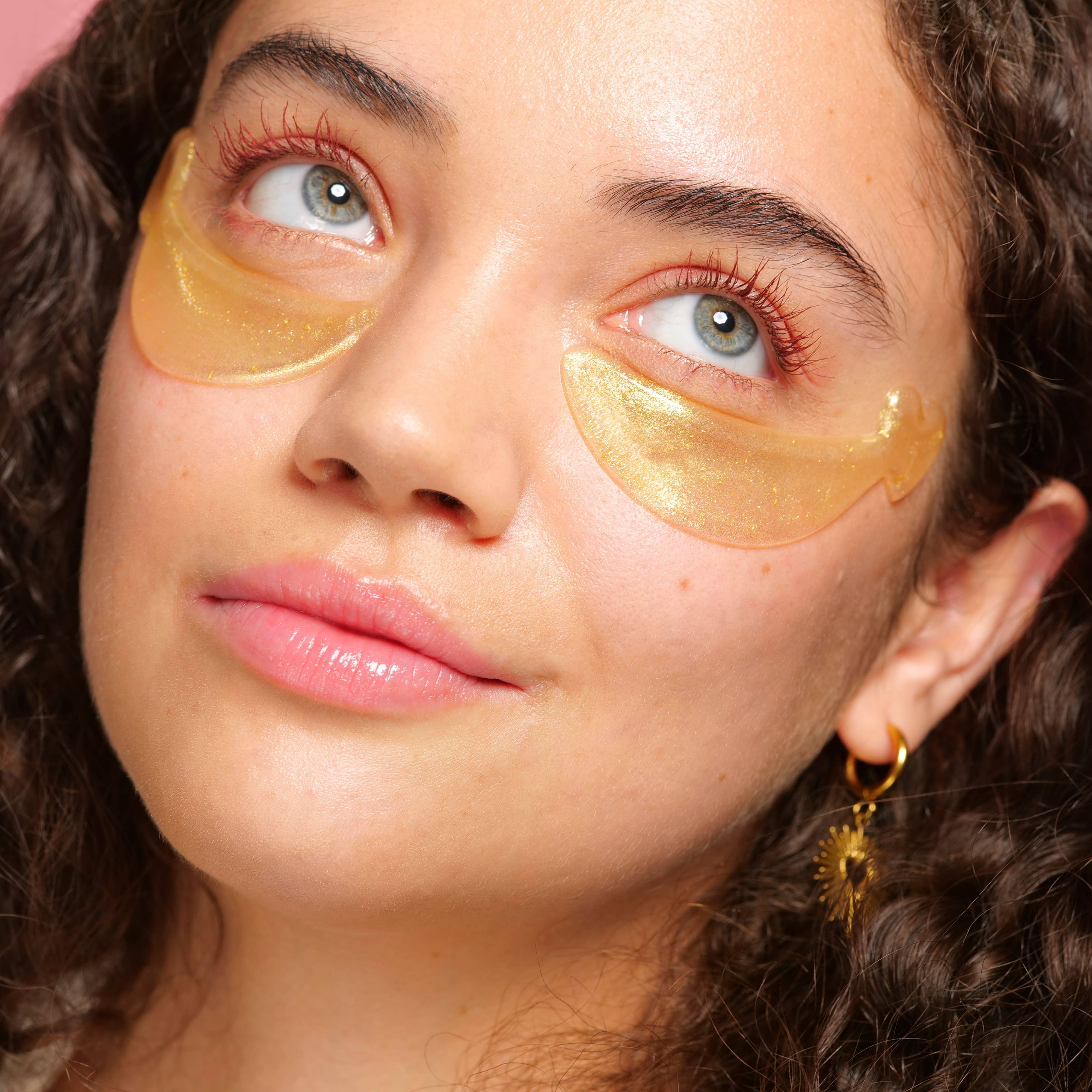 Woman with curly hair wears gold under-eye patches and gold earrings, looking contemplative. The tone is relaxed, with a skincare focus.