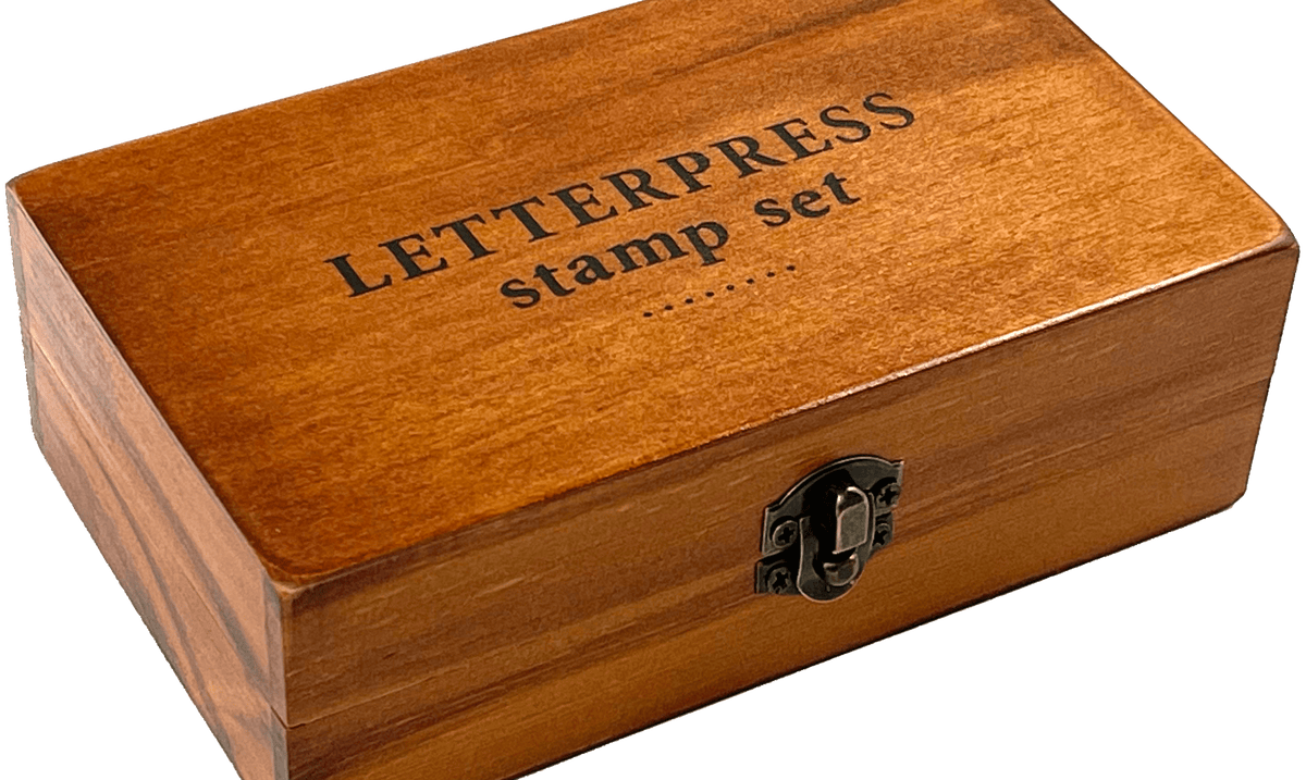 A wooden box labeled "LETTERPRESS stamp set" with a metal latch, resting on a transparent background. The box has a warm, polished finish.