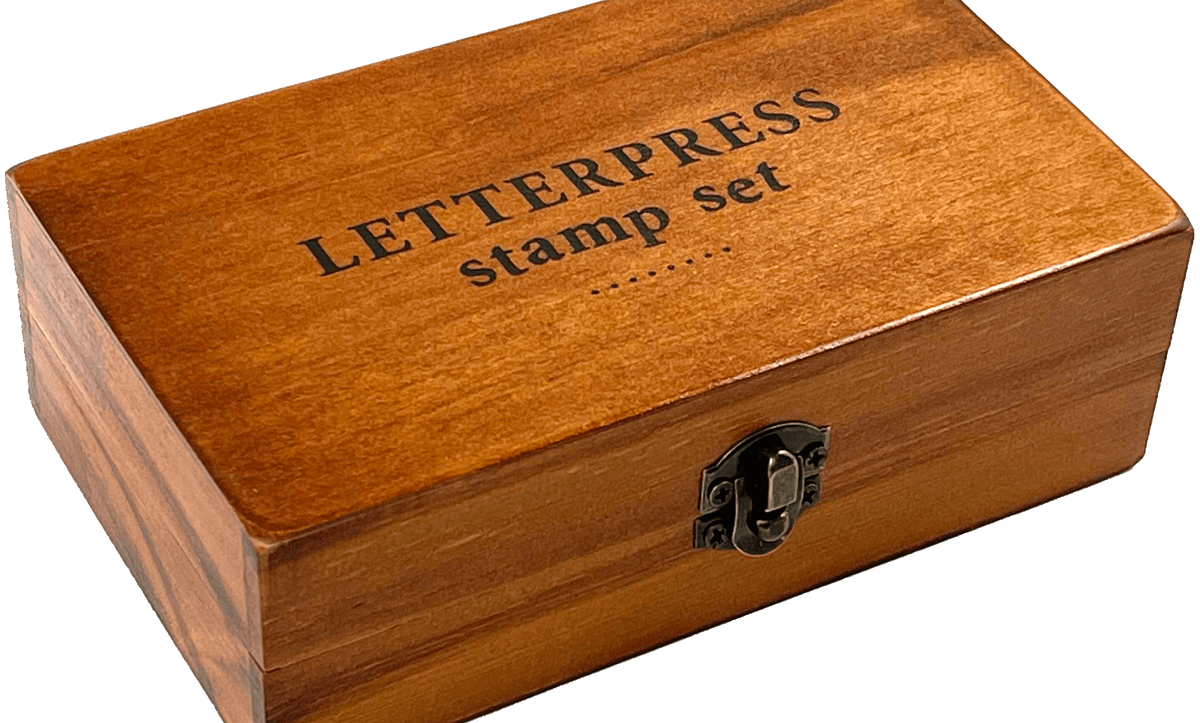A wooden box labeled "LETTERPRESS stamp set" with a metal latch, resting on a transparent background. The box has a warm, polished finish.