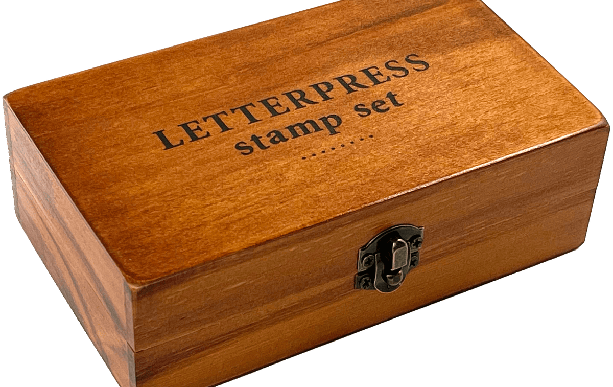 A wooden box labeled "LETTERPRESS stamp set" with a metal latch, resting on a transparent background. The box has a warm, polished finish.
