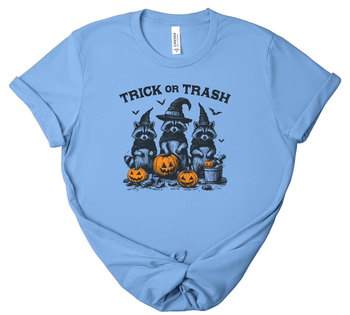 A light blue short-sleeve t-shirt with a 'Trick or Trash' Halloween graphic featuring raccoons in witch hats and jack-o'-lanterns, displayed flat against a white background.