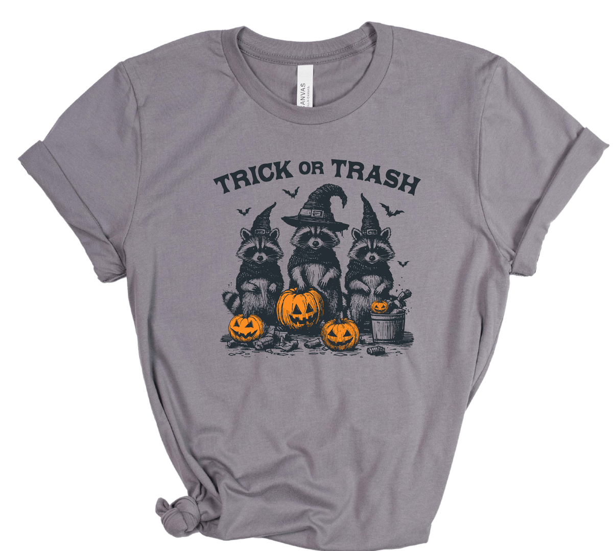 A heather gray short-sleeve t-shirt with a distressed graphic of three raccoons dressed as witches wearing pointed hats, standing behind three jack-o'-lantern pumpkins. The text 'TRICK OR TRASH' is printed above the graphic.