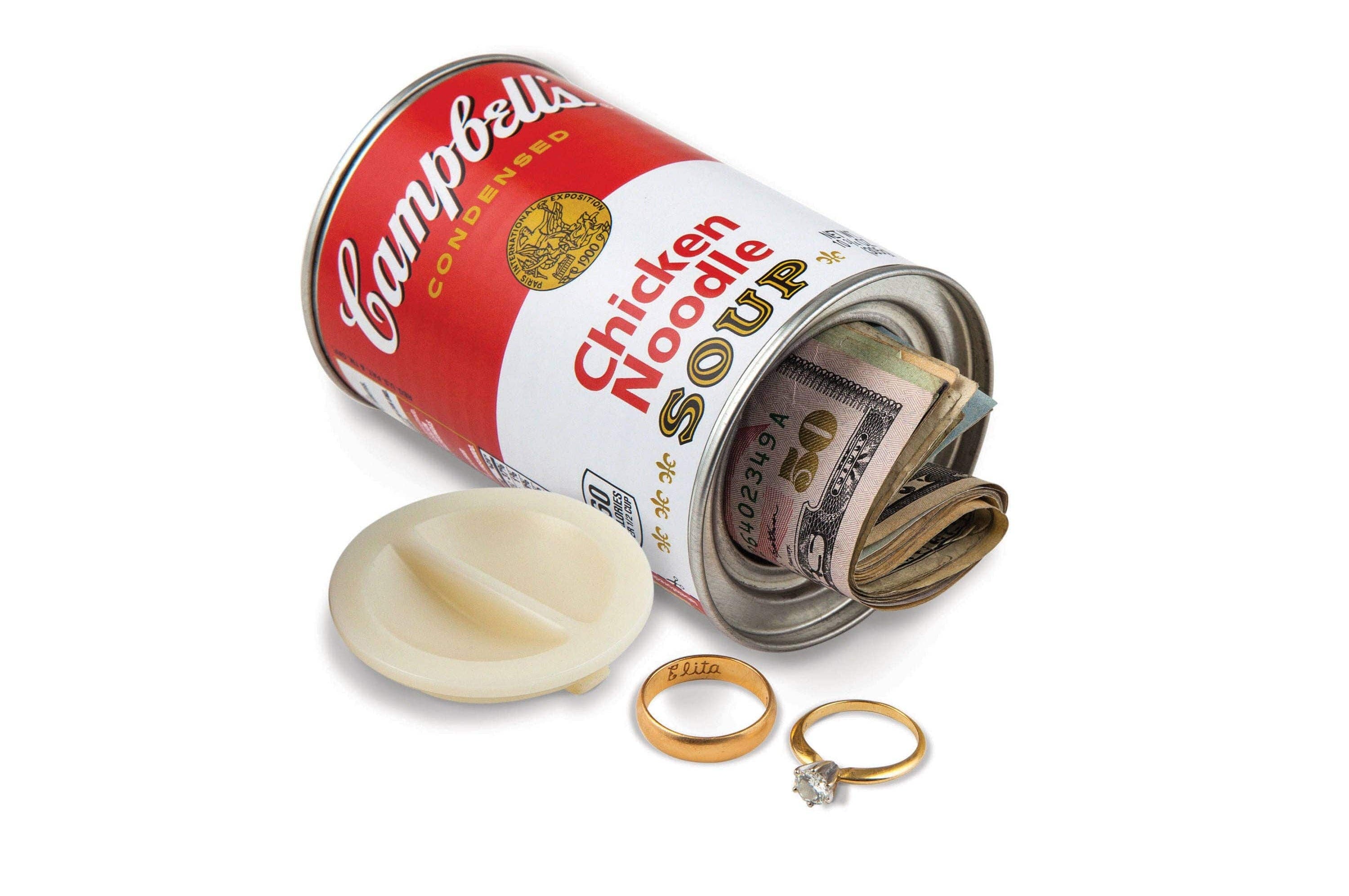 A Campbell's Chicken Noodle Soup can, with its lid removed, reveals rolled U.S. dollar bills inside. Two rings are placed nearby, evoking a sense of hidden treasure.