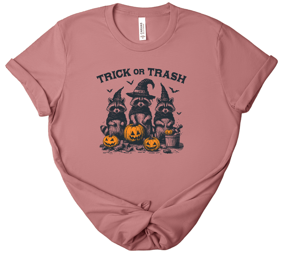 A dusty rose short-sleeve t-shirt with a 'Trick or Trash' Halloween graphic featuring three raccoons in witch hats and jack-o'-lanterns, laid flat on a white background.
