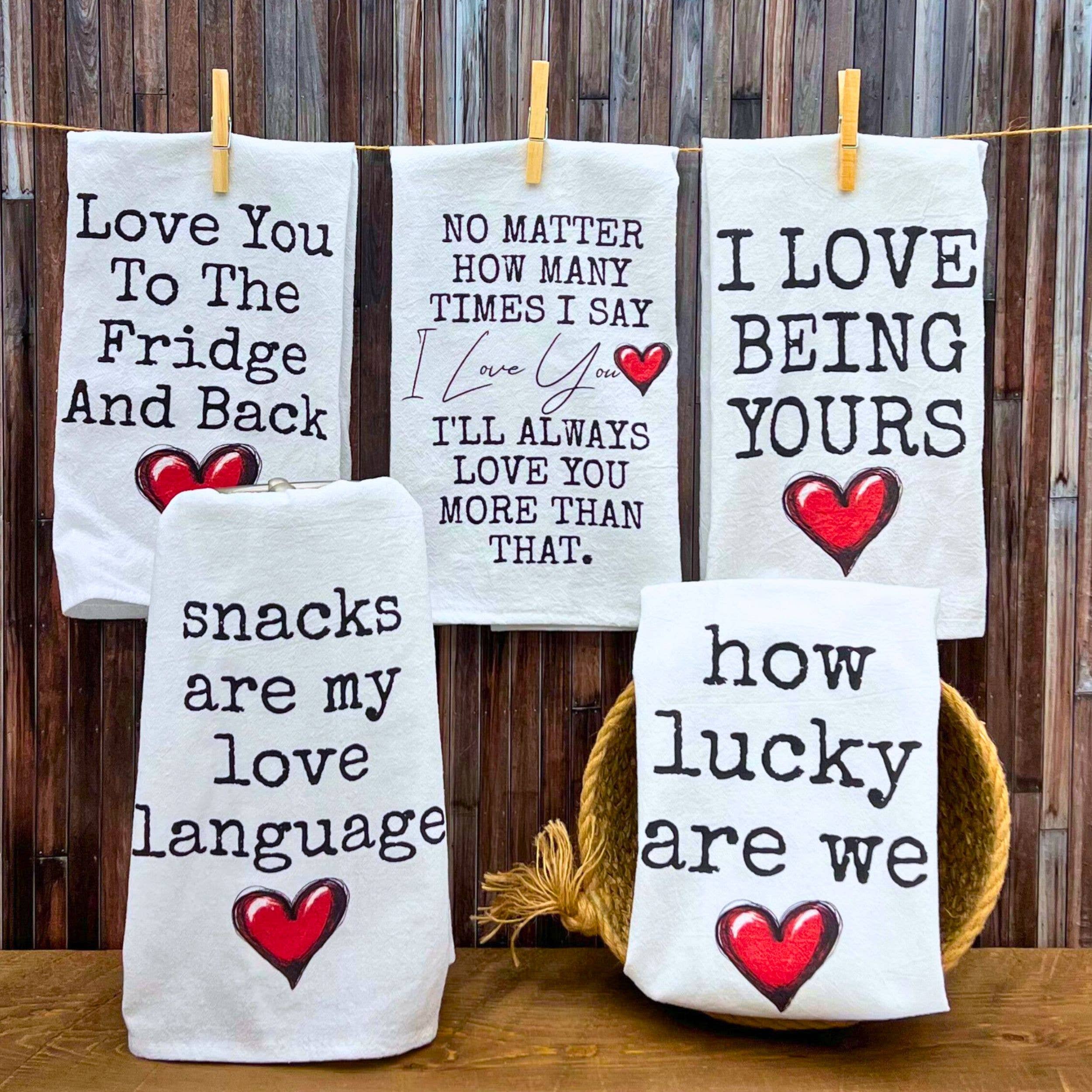 Five towels with playful love quotes are displayed against a wooden wall. Each towel features bold text and red heart graphics, conveying warmth and affection.