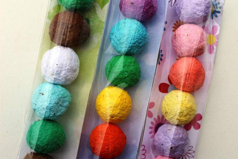 Three packs of colorful seed balls in transparent plastic. Each pack has balls lined in vibrant hues, such as green, blue, pink, and brown, with floral backgrounds.