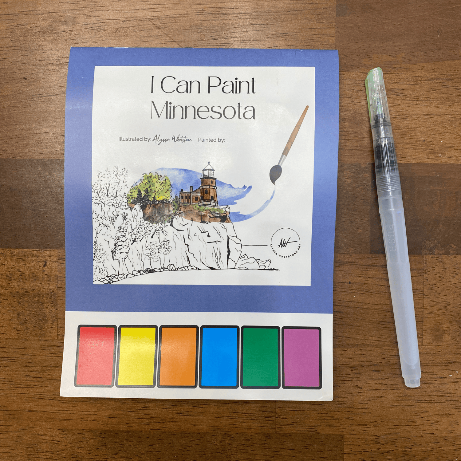 Coloring book titled 'I Can Paint Minnesota' with watercolor pan set and pen on a wooden surface