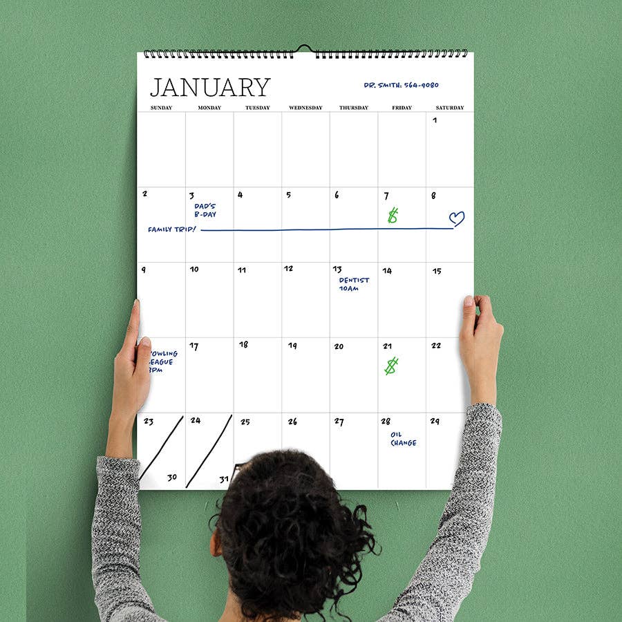A person hangs a large January calendar on a green wall. Key dates include a family trip, dad's birthday, a dentist appointment, and an oil change.