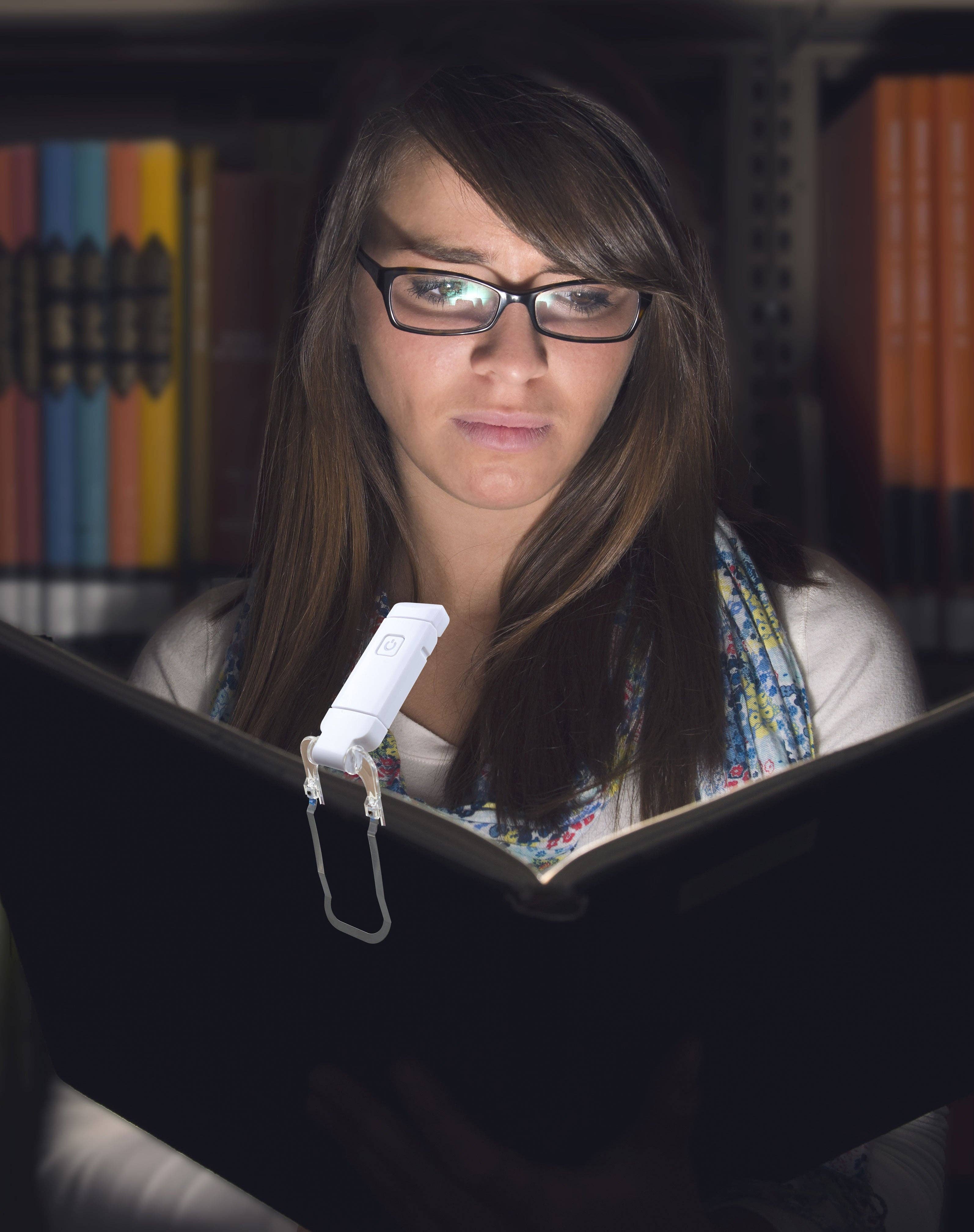 A young woman with glasses reads a book in a dimly lit library, illuminated by a clip-on LED light. She appears focused and engaged.