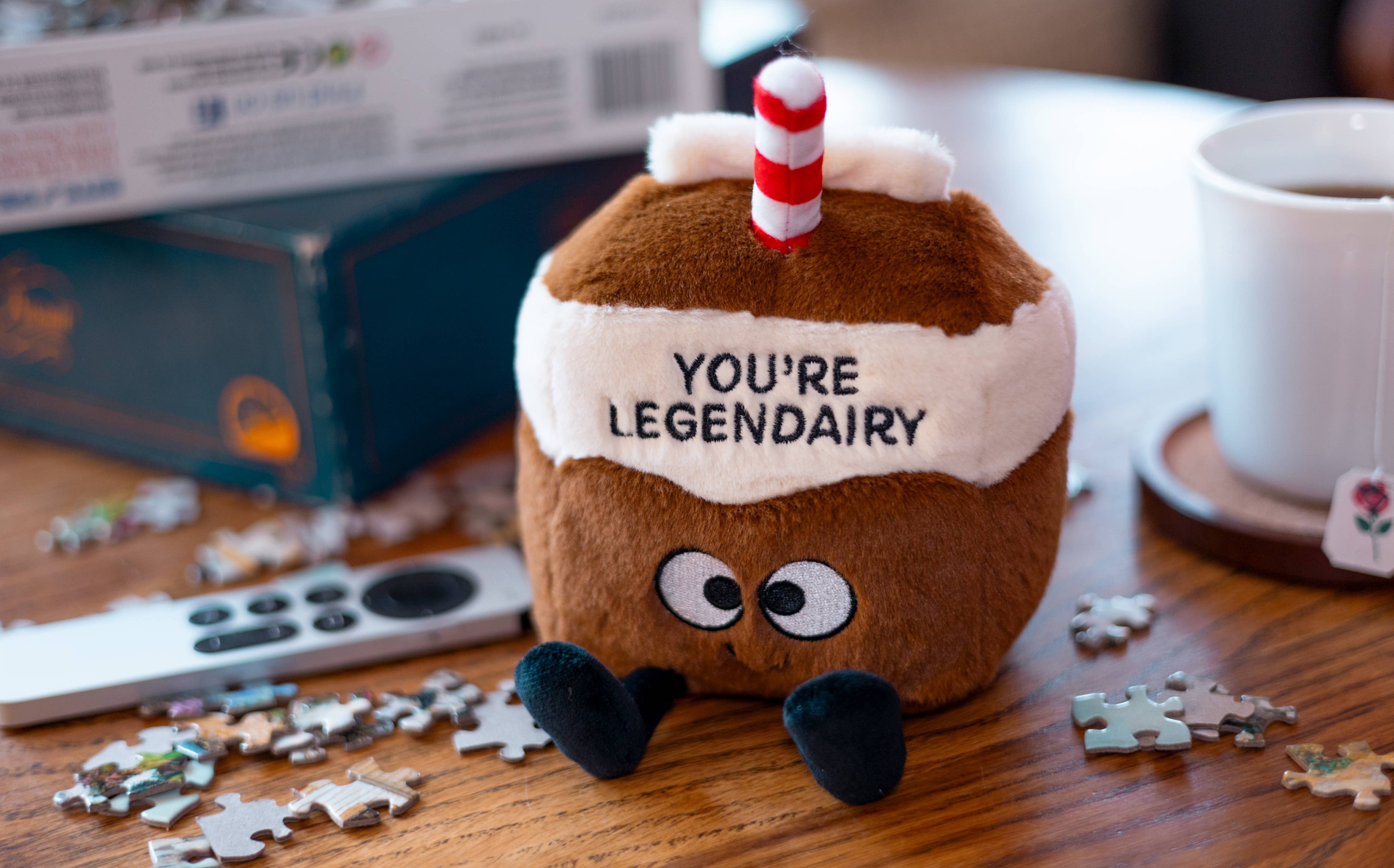 Plush toy resembling a hot chocolate with a straw on a table with puzzle pieces and a mug.