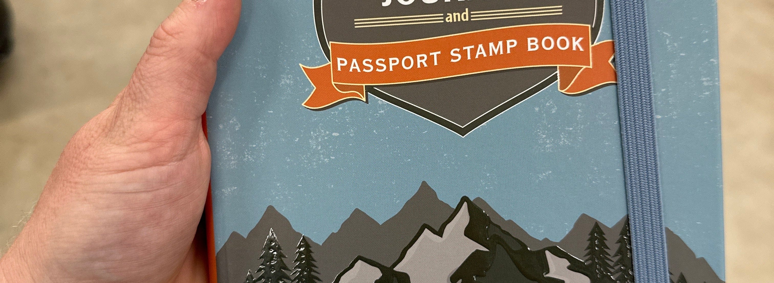 Person holding a "USA National Parks Journal and Passport Stamp Book" with mountain illustration on the cover. The tone is adventurous and inviting.