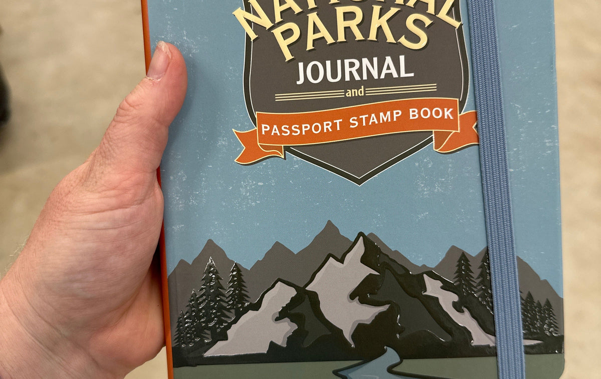 Person holding a "USA National Parks Journal and Passport Stamp Book" with mountain illustration on the cover. The tone is adventurous and inviting.