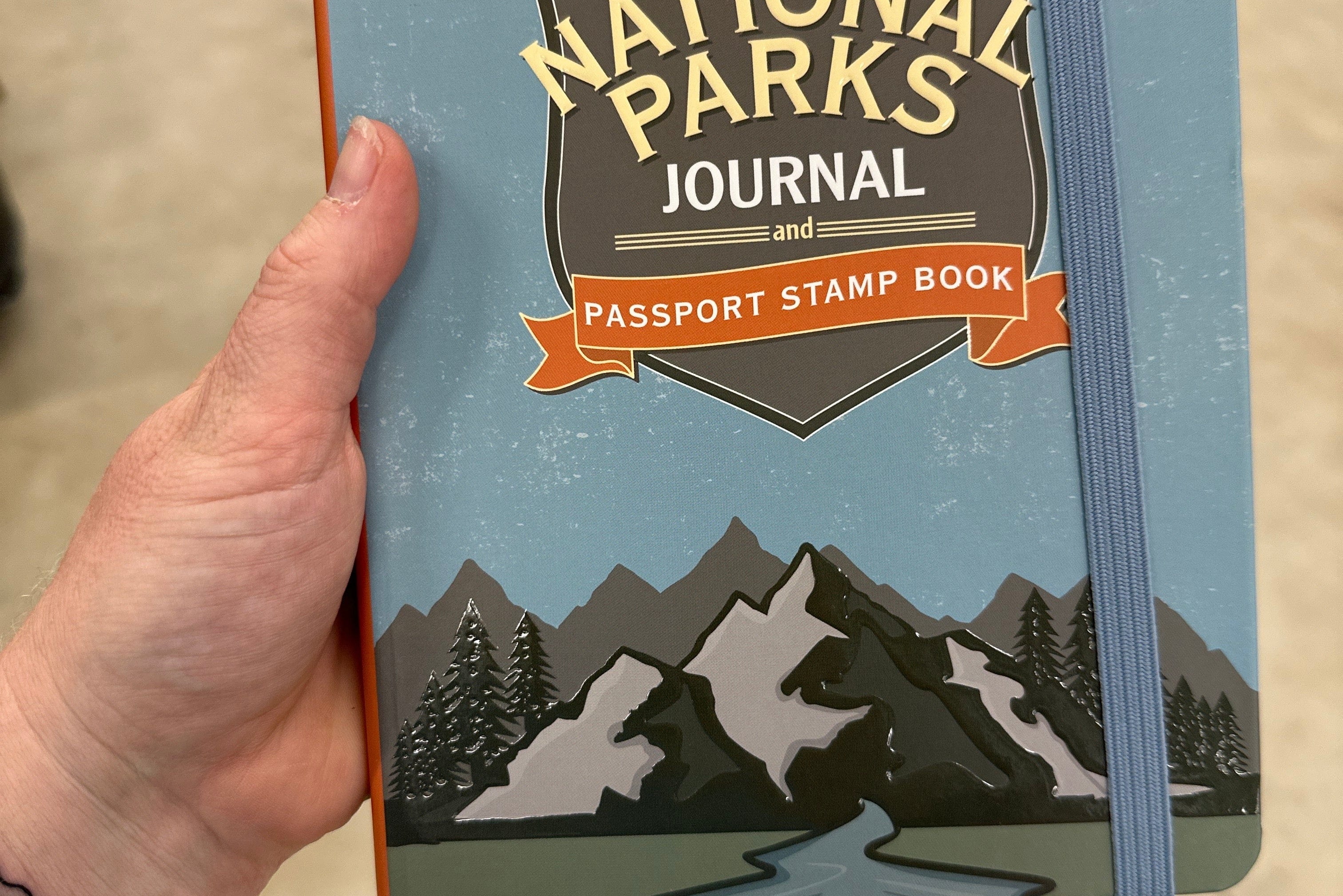 Person holding a "USA National Parks Journal and Passport Stamp Book" with mountain illustration on the cover. The tone is adventurous and inviting.