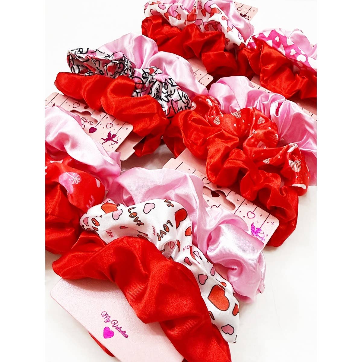 A collection of vibrant scrunchies in red and pink hues, some patterned with hearts and words, displayed on love-themed packaging with a playful, romantic vibe.