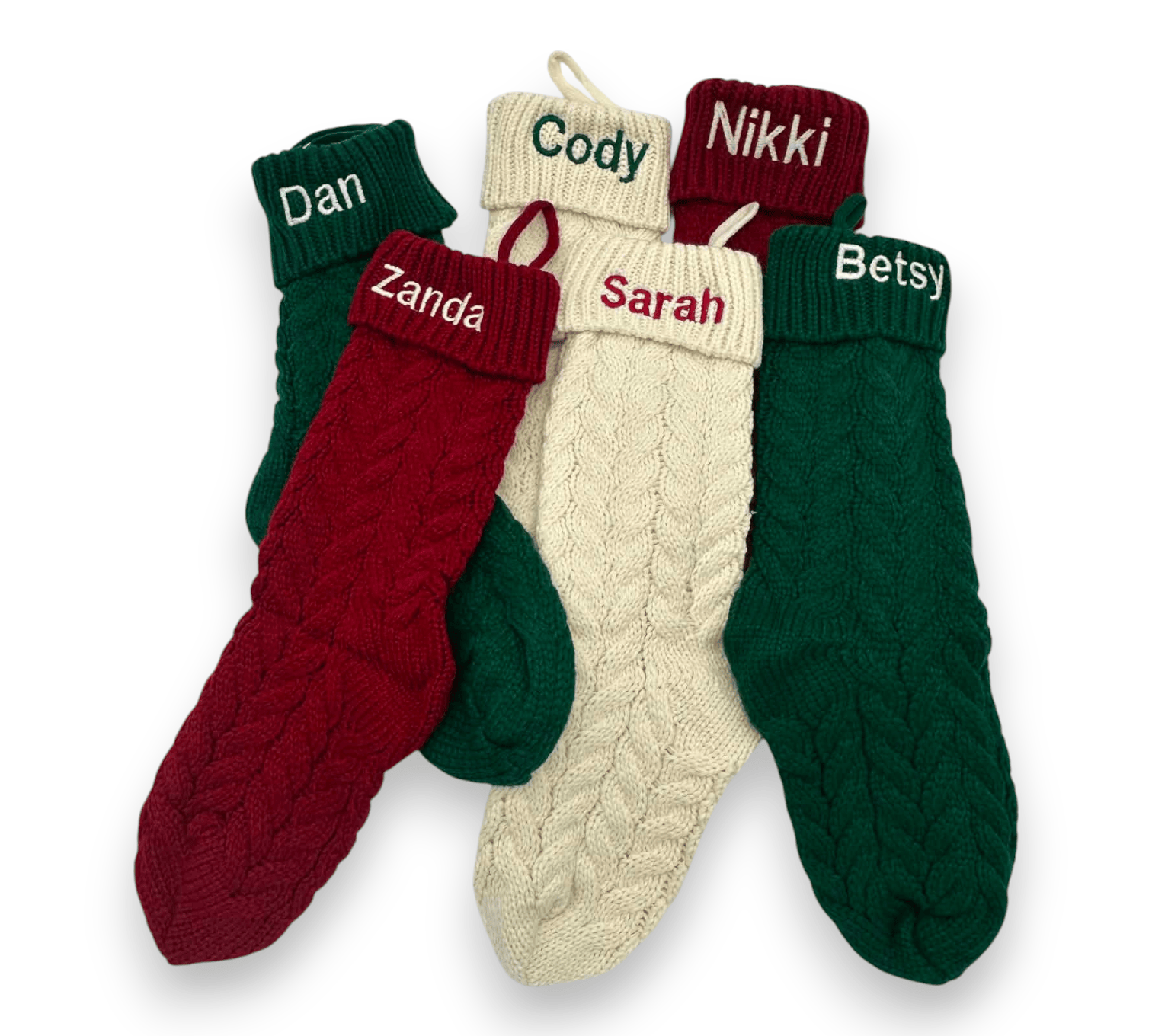 Four personalized cable-knit Christmas socks in green, red, and cream, arranged in a fan shape on a white background. Each sock has a custom name embroidered on the red cuff: Dan, Zanda, Cody, and Sarah, with Betsy also visible on another cuff.