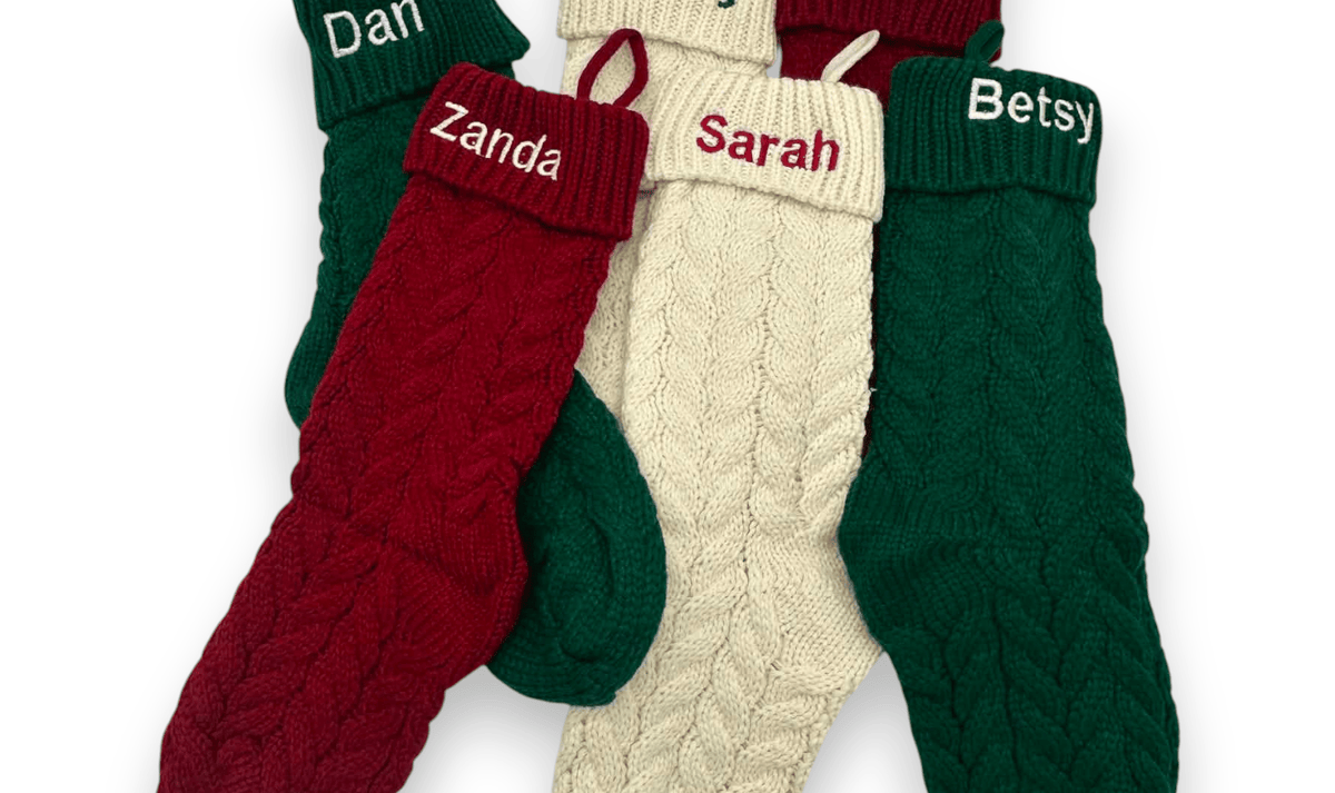 Four personalized cable-knit Christmas socks in green, red, and cream, arranged in a fan shape on a white background. Each sock has a custom name embroidered on the red cuff: Dan, Zanda, Cody, and Sarah, with Betsy also visible on another cuff.