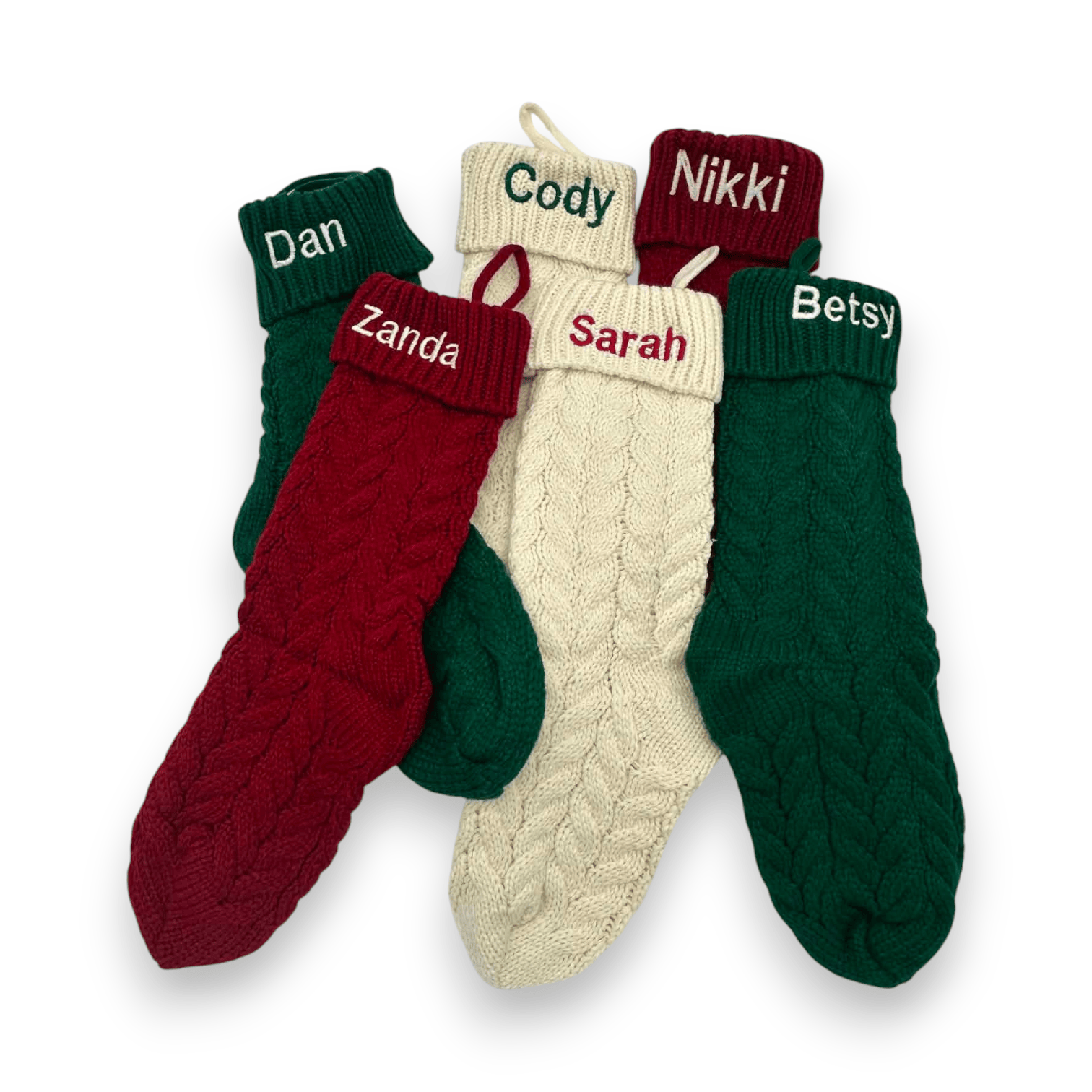 Four personalized cable-knit Christmas socks in green, red, and cream, arranged in a fan shape on a white background. Each sock has a custom name embroidered on the red cuff: Dan, Zanda, Cody, and Sarah, with Betsy also visible on another cuff.