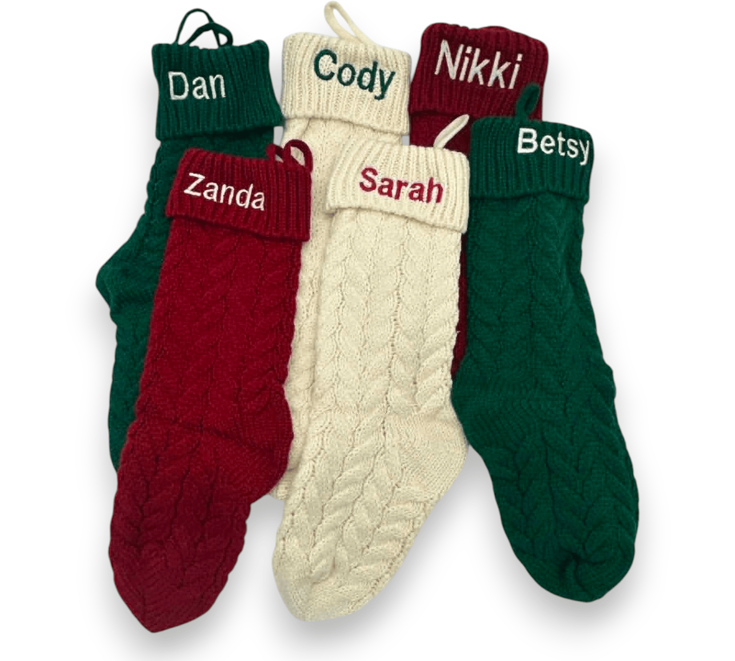 Four personalized cable-knit Christmas socks in green, cream, and burgundy, arranged together on a white background. Each sock has a different name embroidered on the cuff: Dan, Cody, Nikki, and Betsy, with Zanda also visible on a burgundy sock.