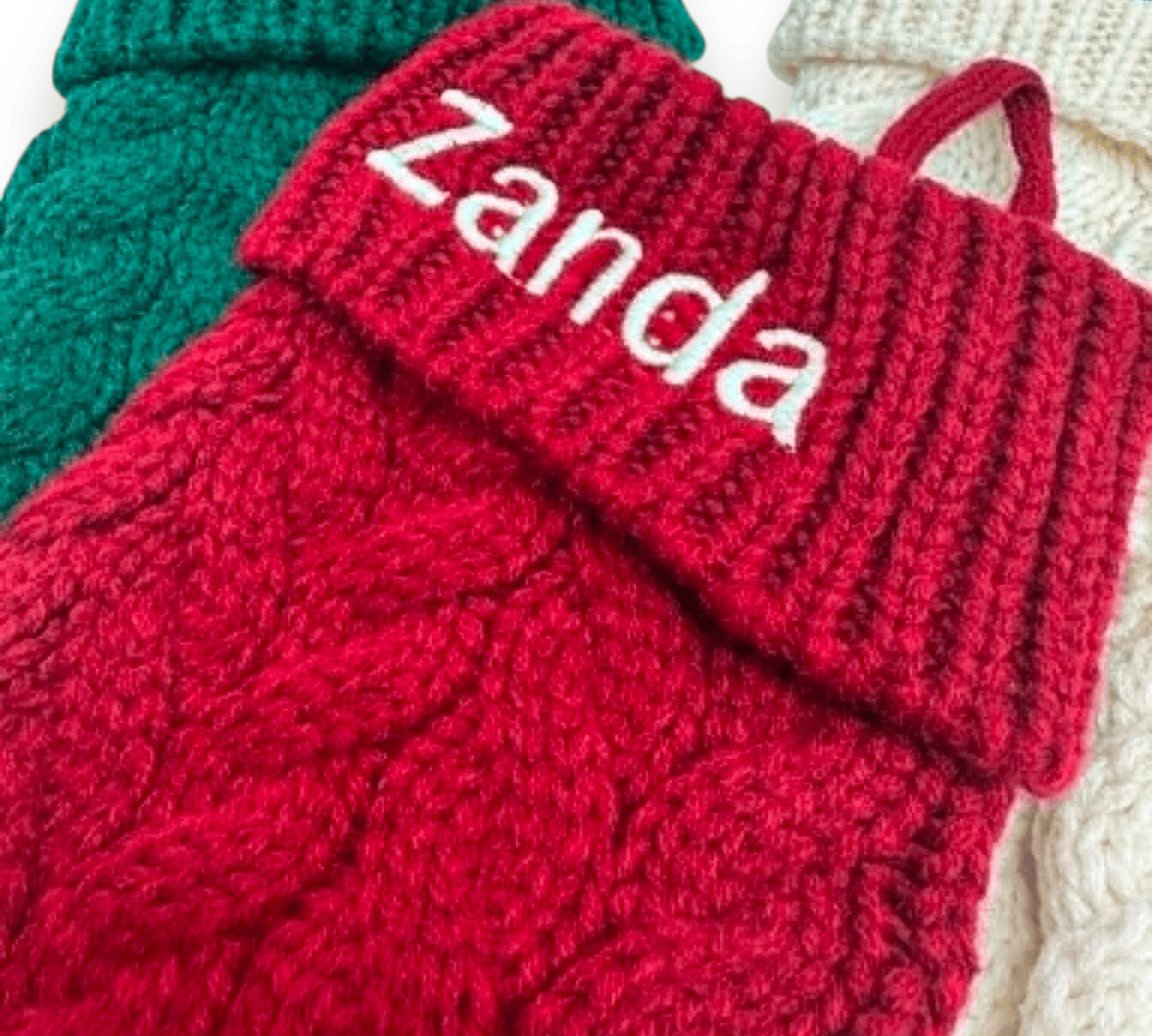 A close-up of a red cable-knit sock holder with a folded cuff embroidered with the name 'Zanda' in white. Other knitted holders in green and cream are partially visible in the background.