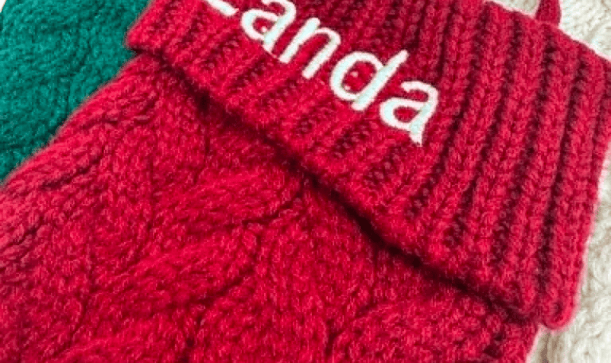 A close-up of a red cable-knit sock holder with a folded cuff embroidered with the name 'Zanda' in white. Other knitted holders in green and cream are partially visible in the background.