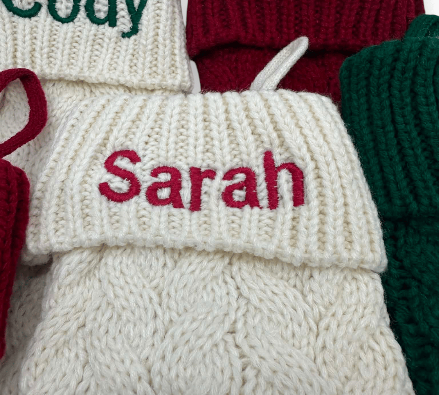 Close-up of a cream knit Christmas stocking with the name 'Sarah' embroidered in red across the cuff, surrounded by other knitted stockings in red, green, and cream.