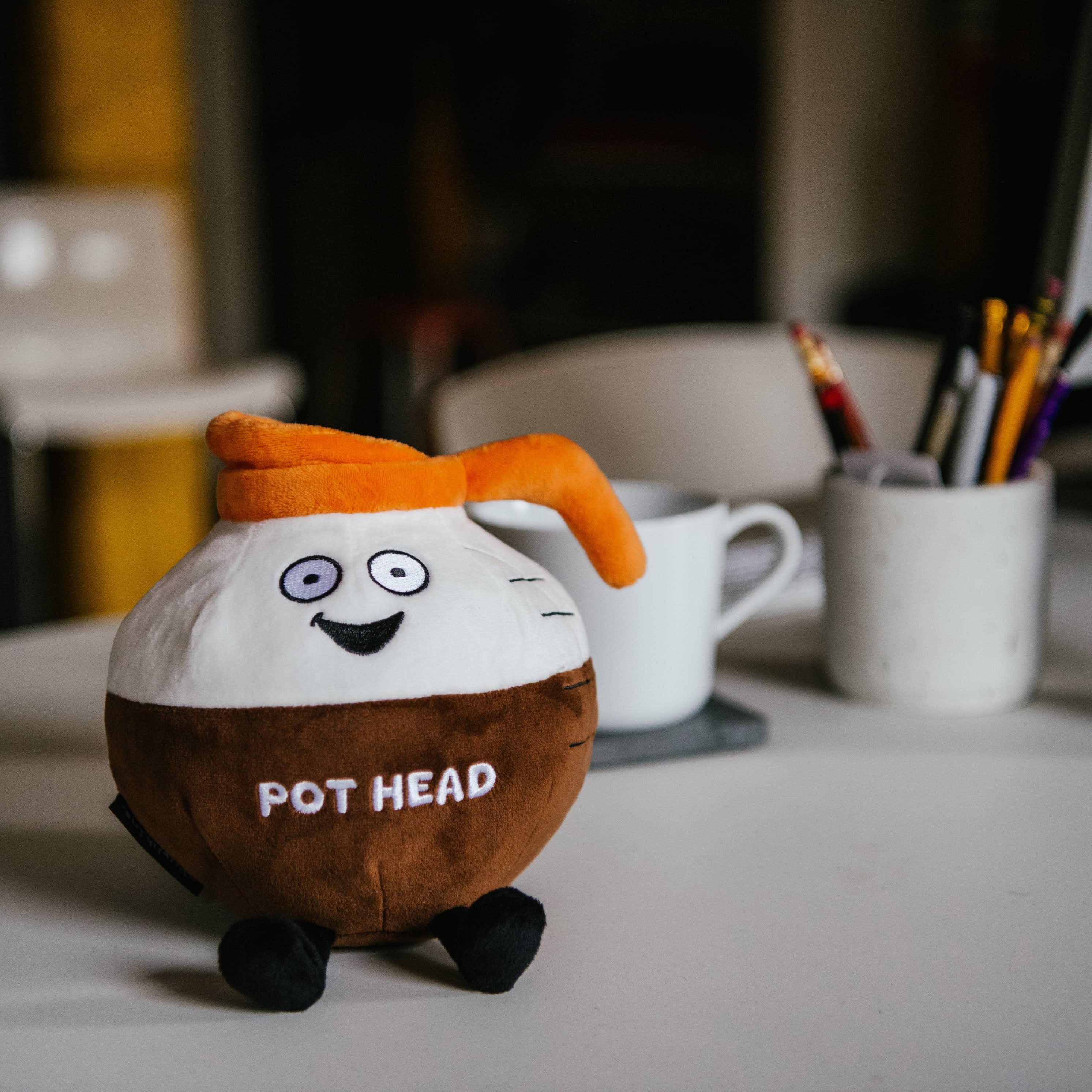 A brown and white coffee pot plushie with an orange pour spout. The white portion has a smiley face, and the brown portion features white text that says "Pot Head." The plushie has black legs sticking out. Plushie is sitting on a desk