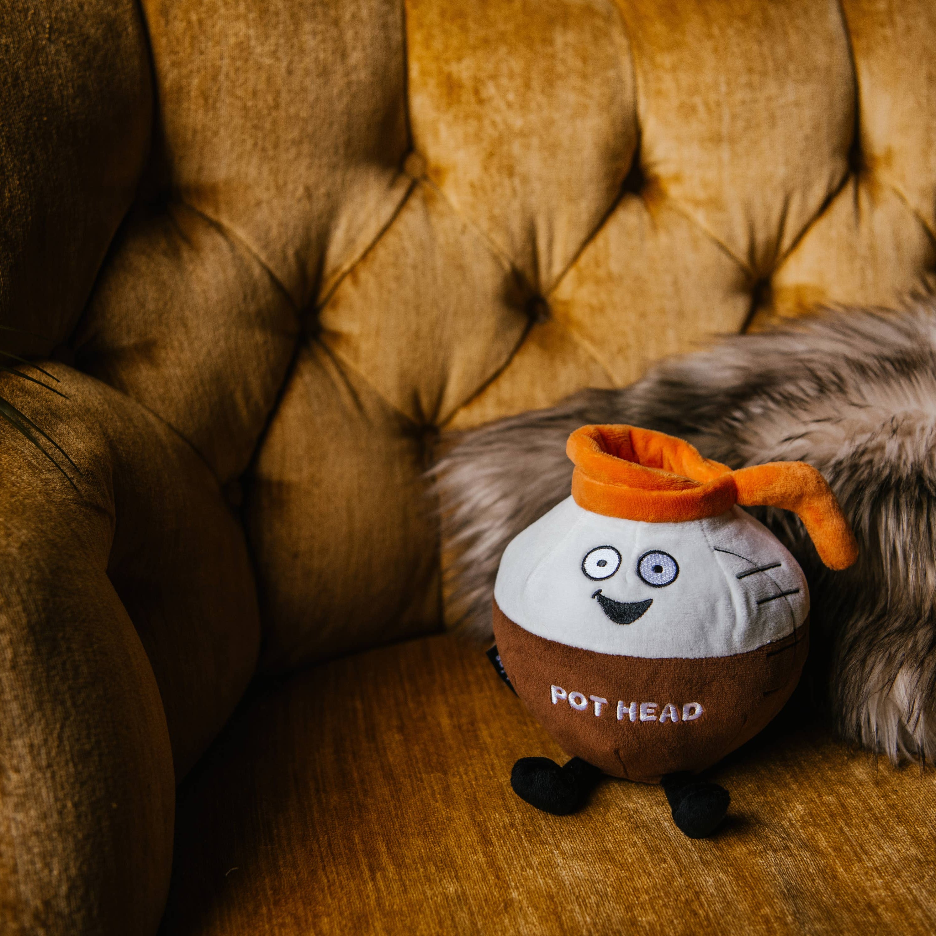 A brown and white coffee pot plushie with an orange pour spout. The white portion has a smiley face, and the brown portion features white text that says "Pot Head." The plushie has black legs sticking out.