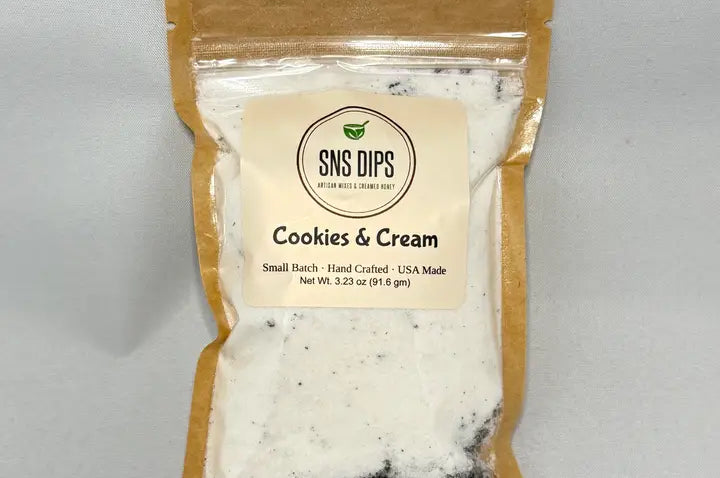 A brown, transparent package labeled "Cookies & Cream" from SNS Dips. It contains a white powder with dark specks, indicating a sweet mix.