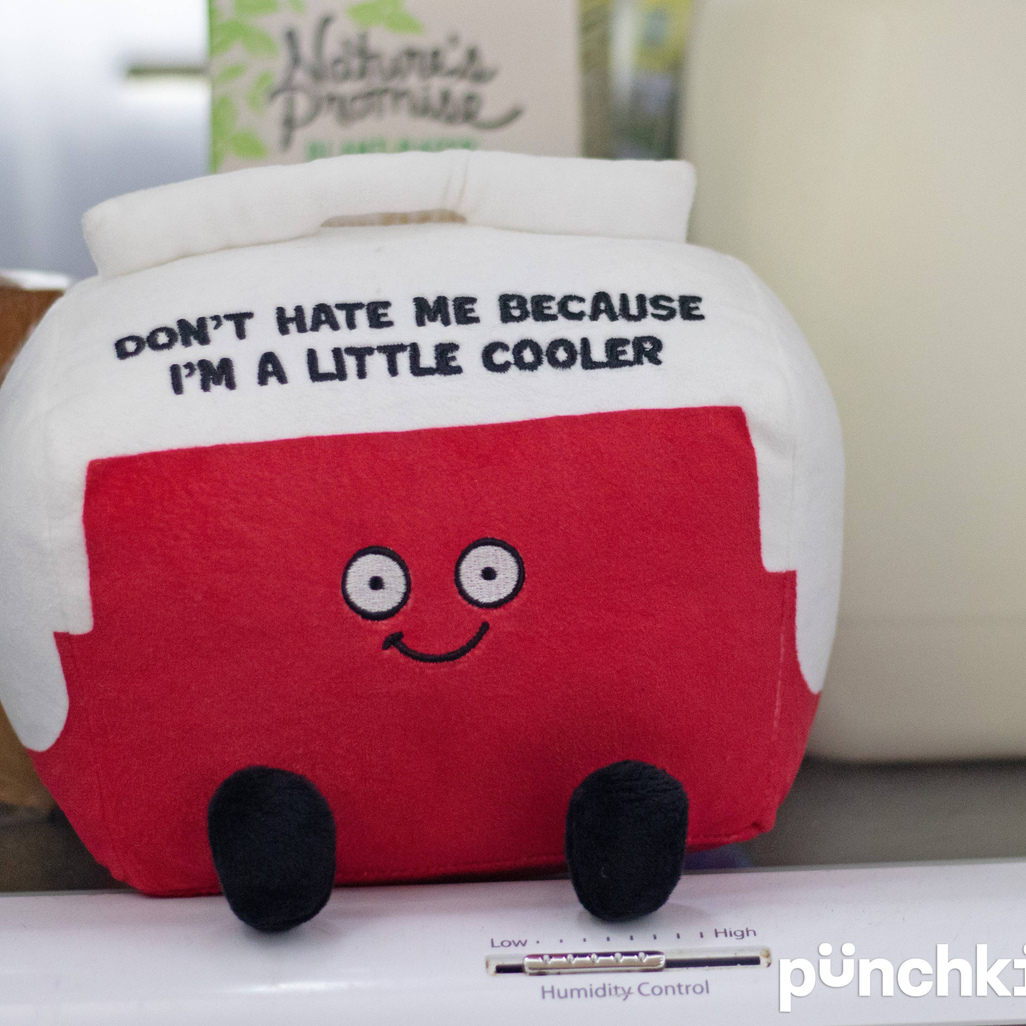 A cooler plushie with a white top and red bottom. The white top features black embroidered text that says, "Don't hate me because I'm a little cooler." The red section has a smiling face, with black legs sticking out from the bottom.