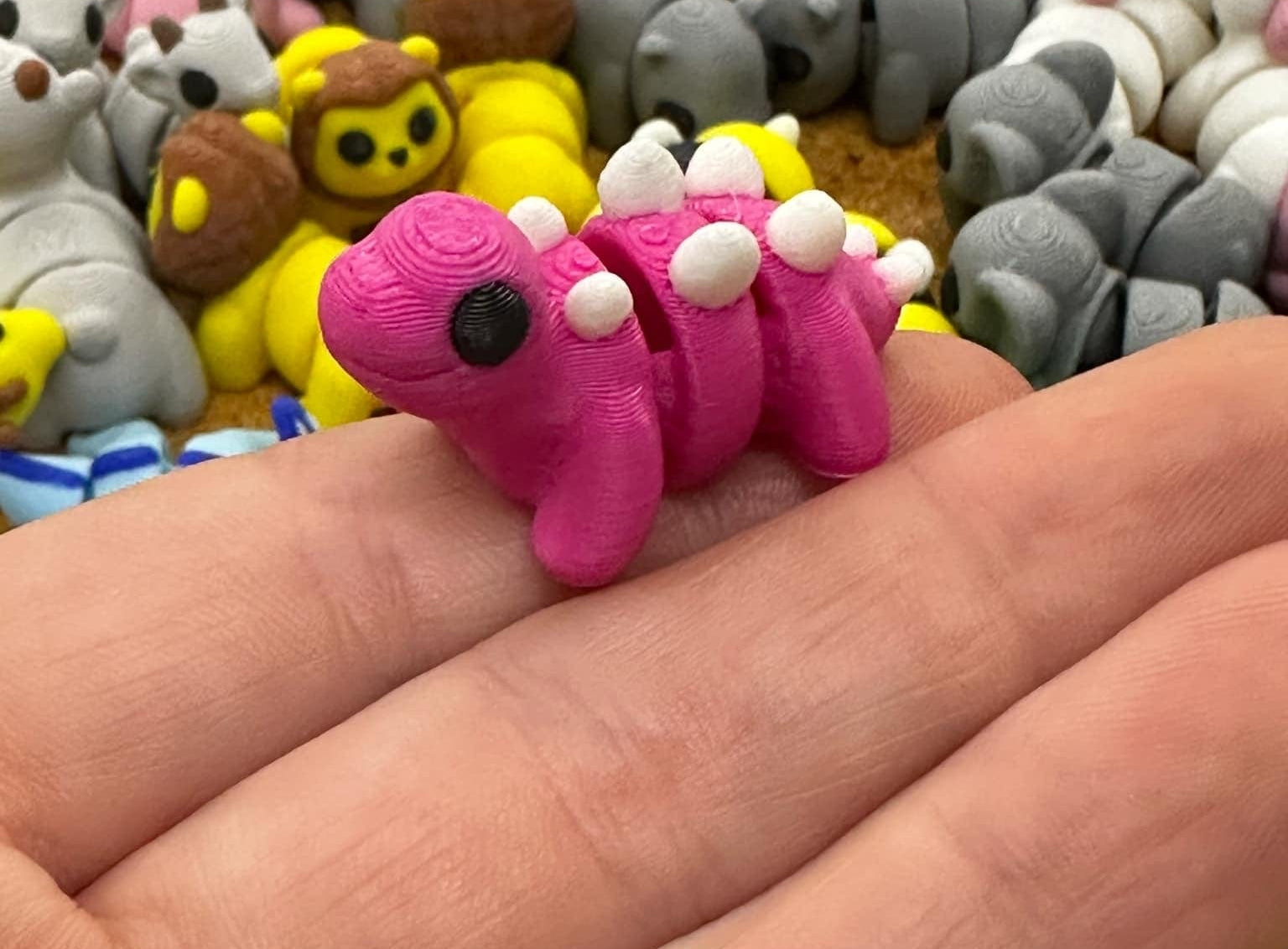 A small, pink, toy dinosaur with white spikes being held between fingers, part of a larger collection of similar mini fidget toys.