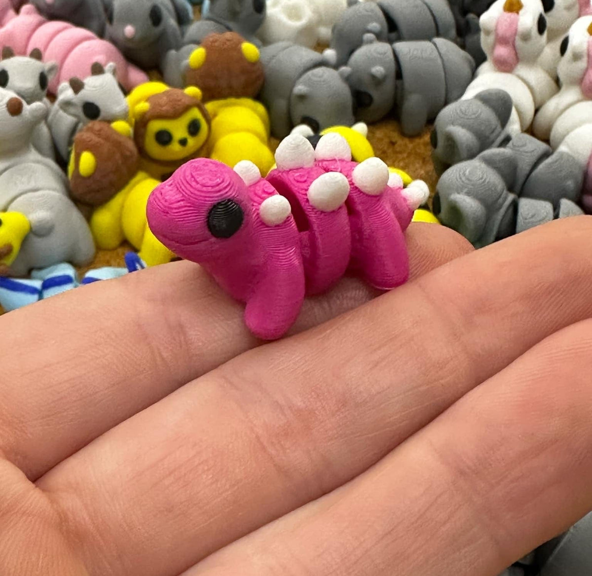 A small, pink, toy dinosaur with white spikes being held between fingers, part of a larger collection of similar mini fidget toys.