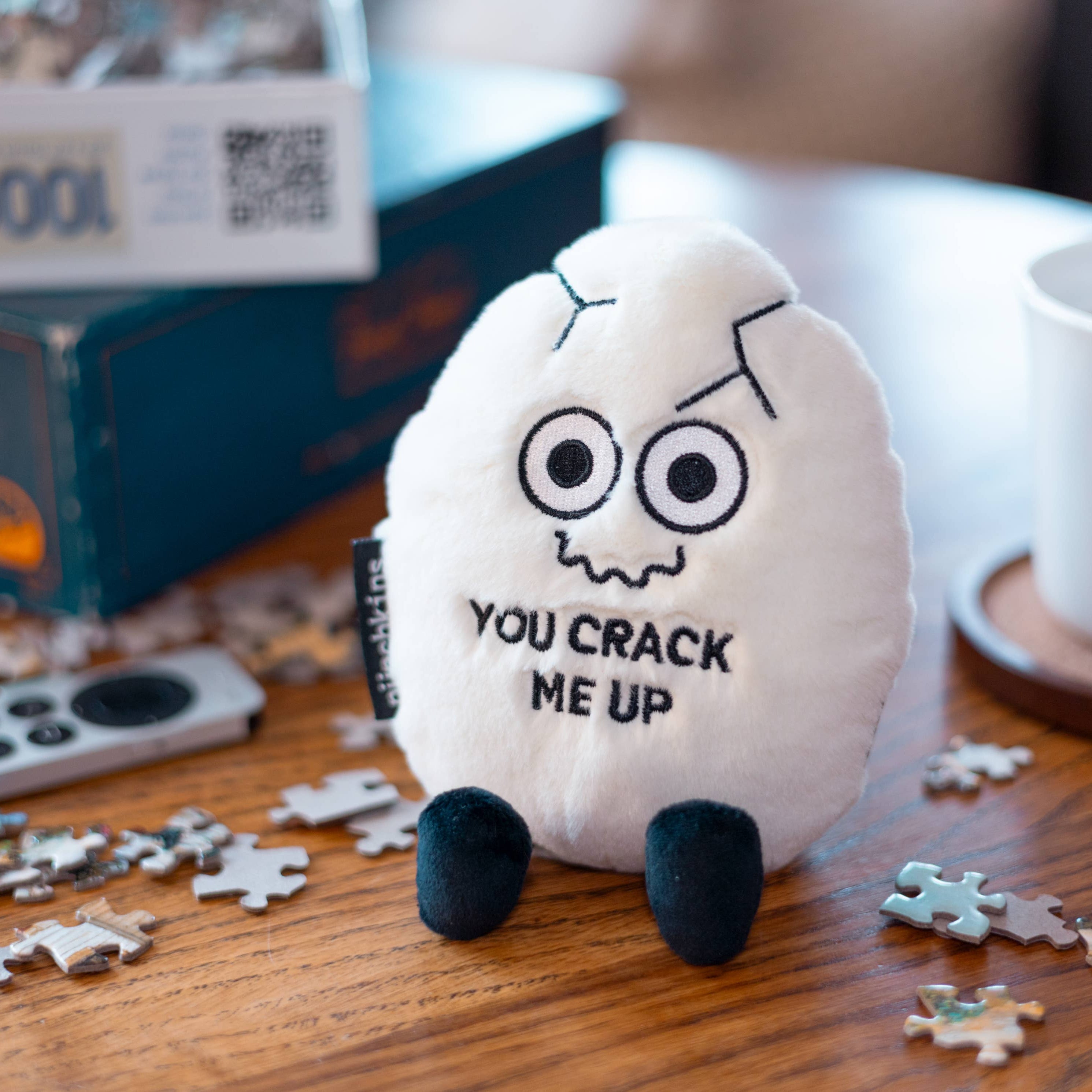 A white cracked egg plush with a squiggly smiley face and black text that says "You crack me up." The plushie has black legs sticking out.