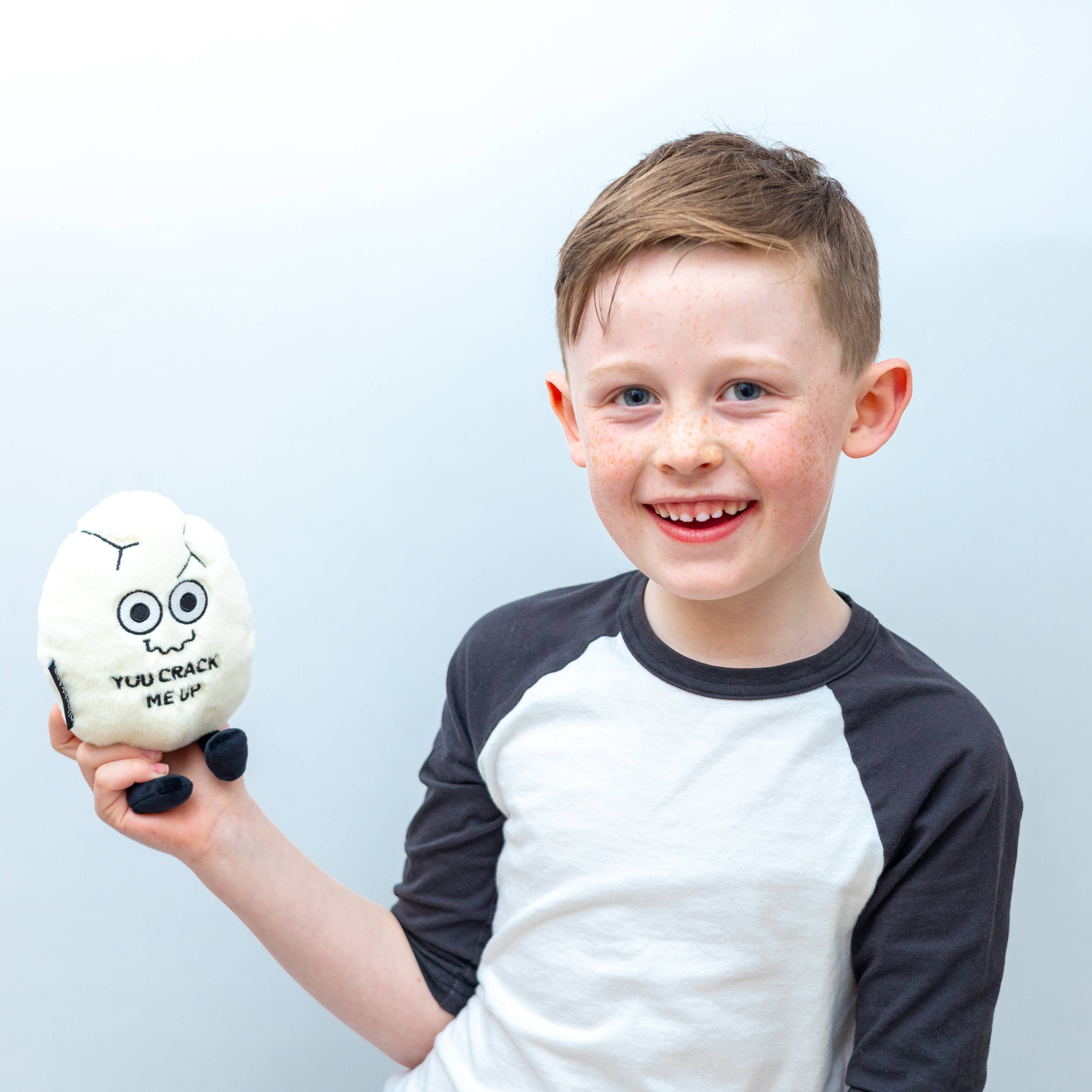 Child holding a white cracked egg plush with a squiggly smiley face and black text that says "You crack me up." The plushie has black legs sticking out.