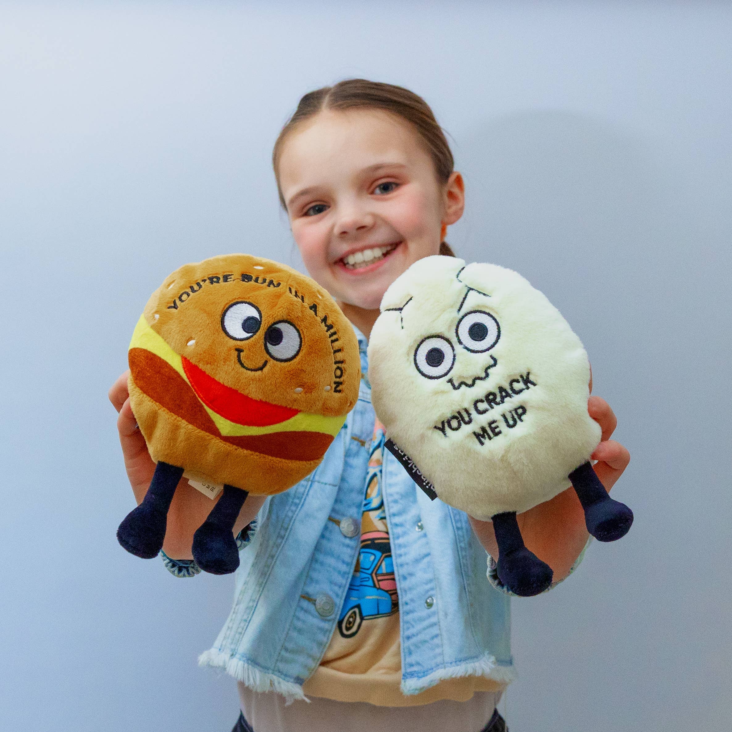 Child holding two plushies. Awhite cracked egg plush with a squiggly smiley face and black text that says "You crack me up." The plushie has black legs sticking out. The other a hamburger plushie