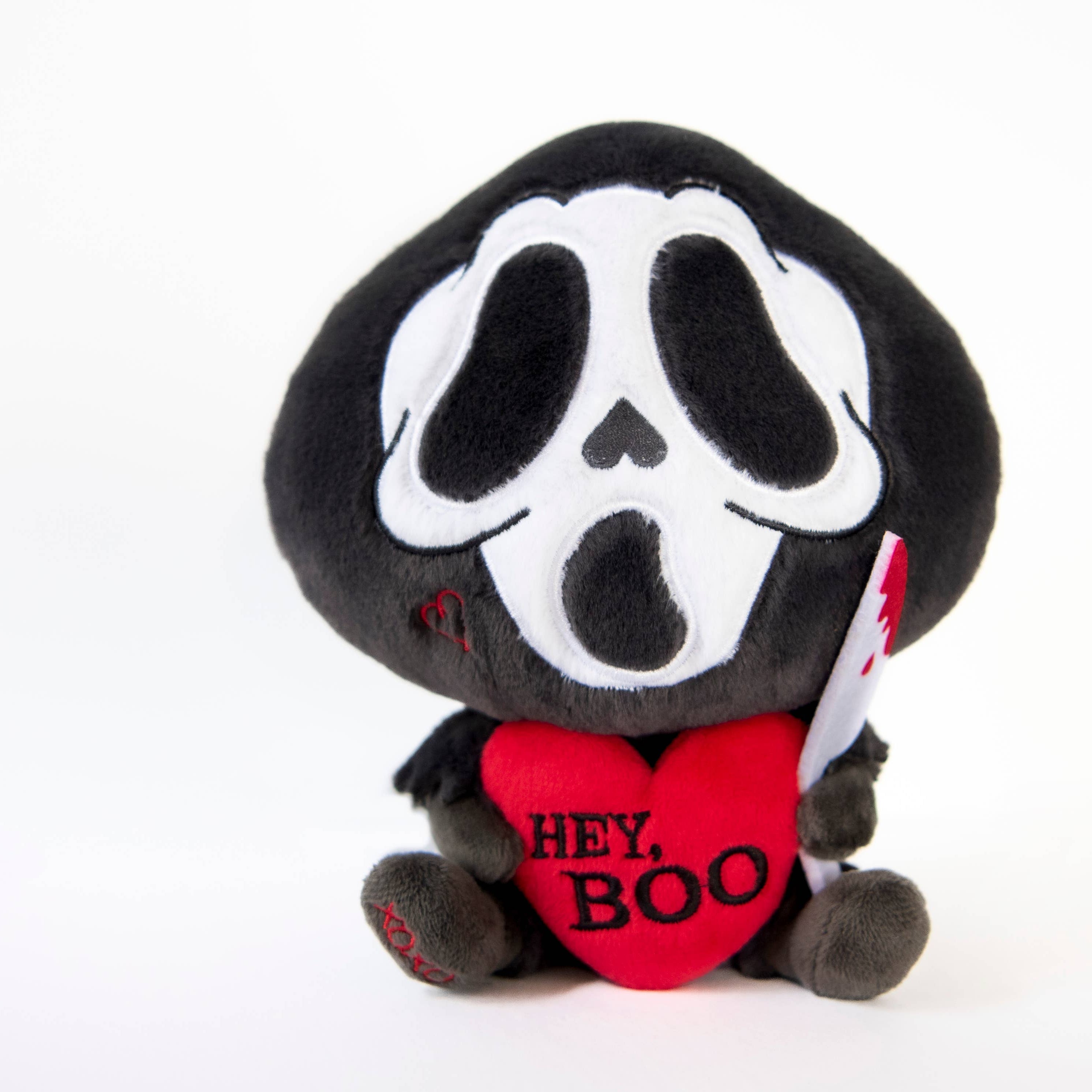 A black and white ghost resembling the Scream mask, holding a bloody knife in one hand and a red heart in the other. The heart has the phrase "Hey, Boo" written in black text.