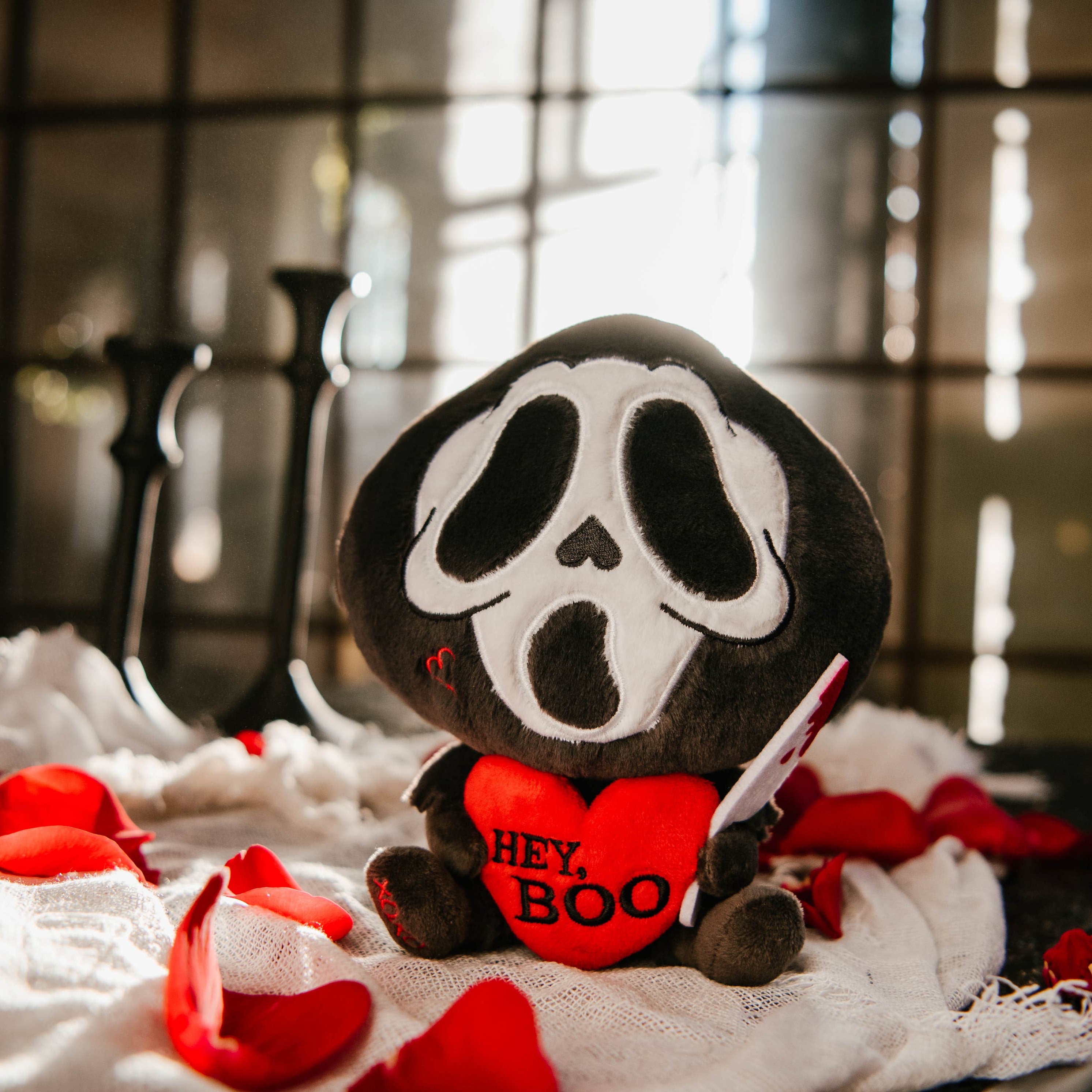 A black and white ghost resembling the Scream mask, holding a bloody knife in one hand and a red heart in the other. The heart has the phrase "Hey, Boo" written in black text.