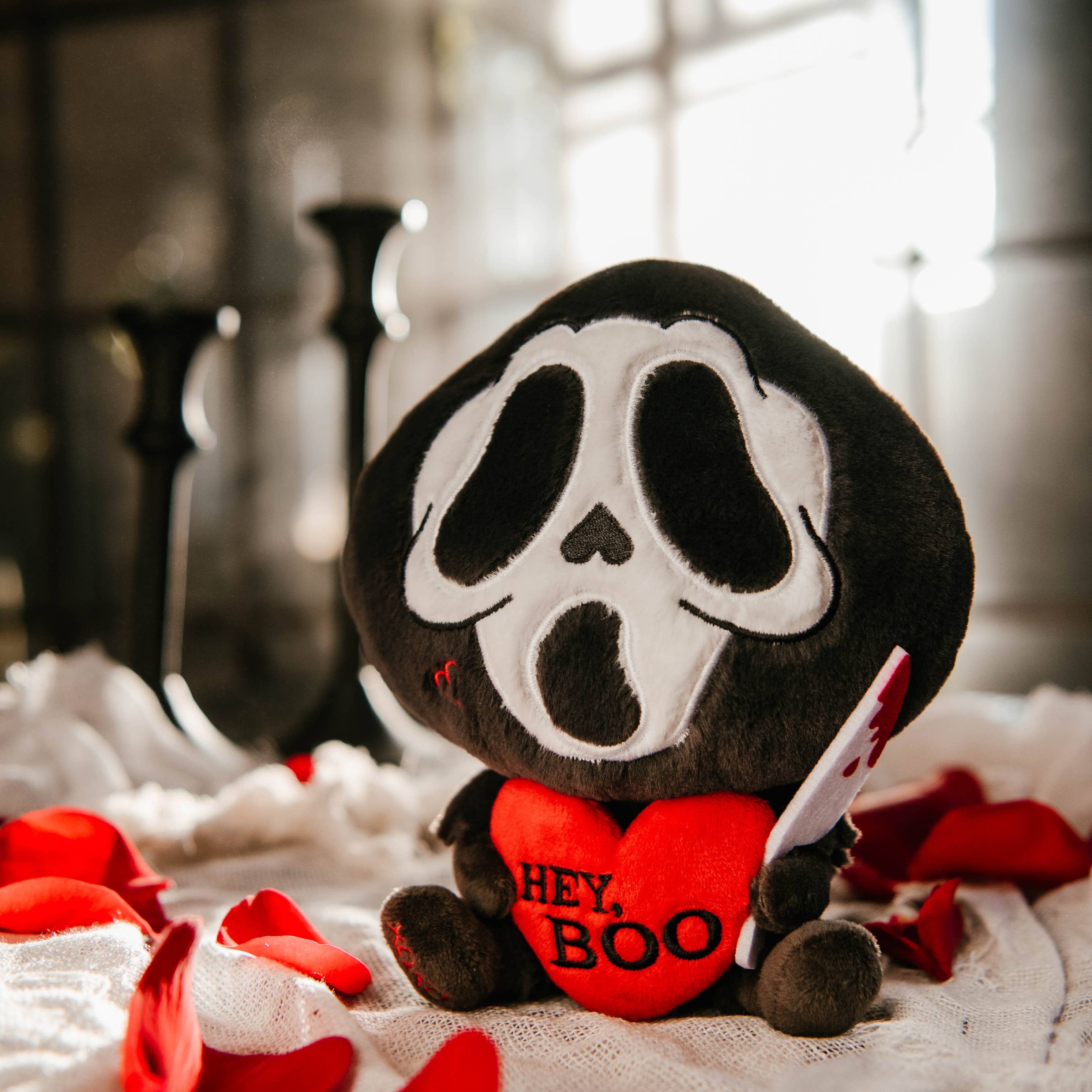 A black and white ghost resembling the Scream mask, holding a bloody knife in one hand and a red heart in the other. The heart has the phrase "Hey, Boo" written in black text.