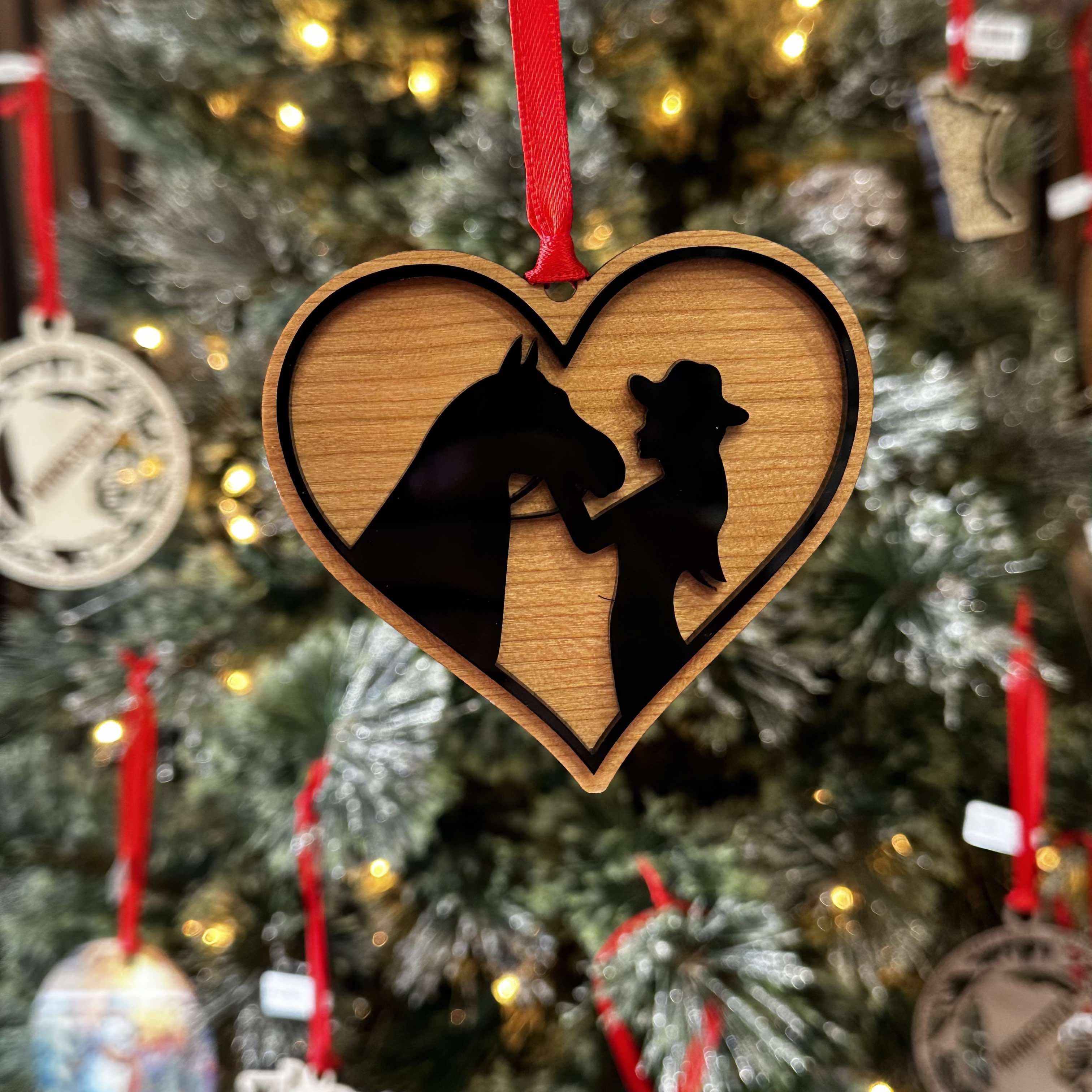A wooden ornament shaped like a heart with a silhouette of a horse and a girl within the heart, hanging from a Christmas tree with other ornaments.