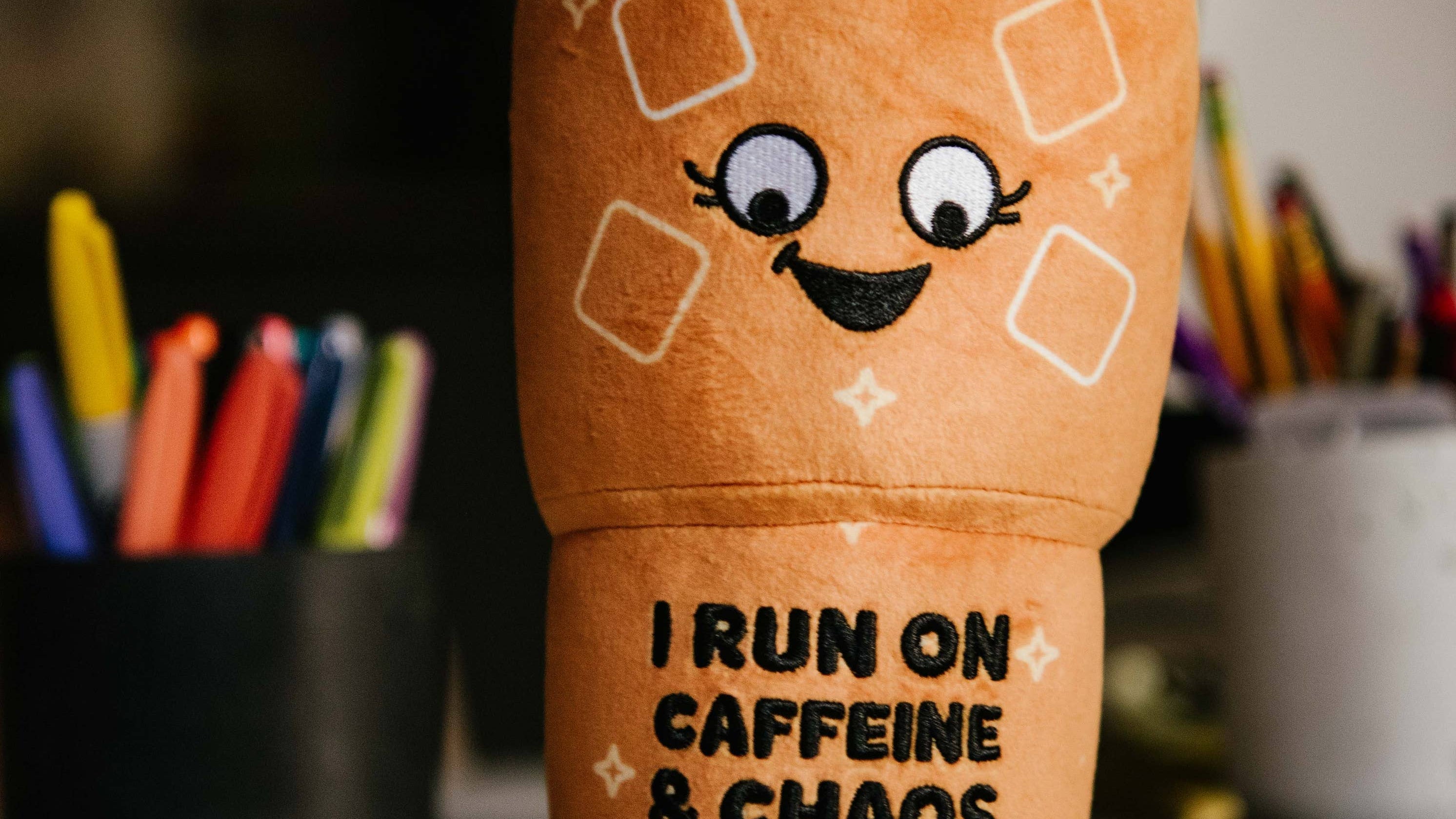 Plushie shaped like a ln iced coffee with a cute face that reads “ I run on caffeine and chaos”