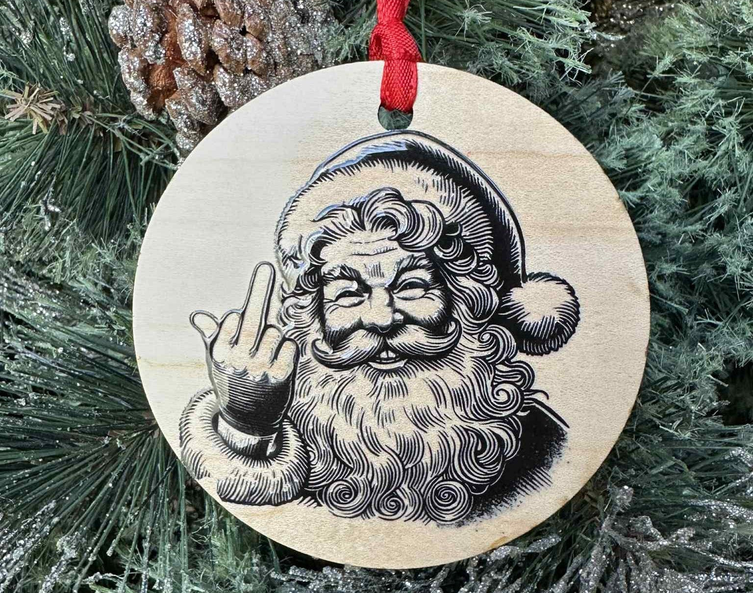Wooden Christmas ornament with a humorous Santa Claus design giving a middle finger, hanging on a tree.